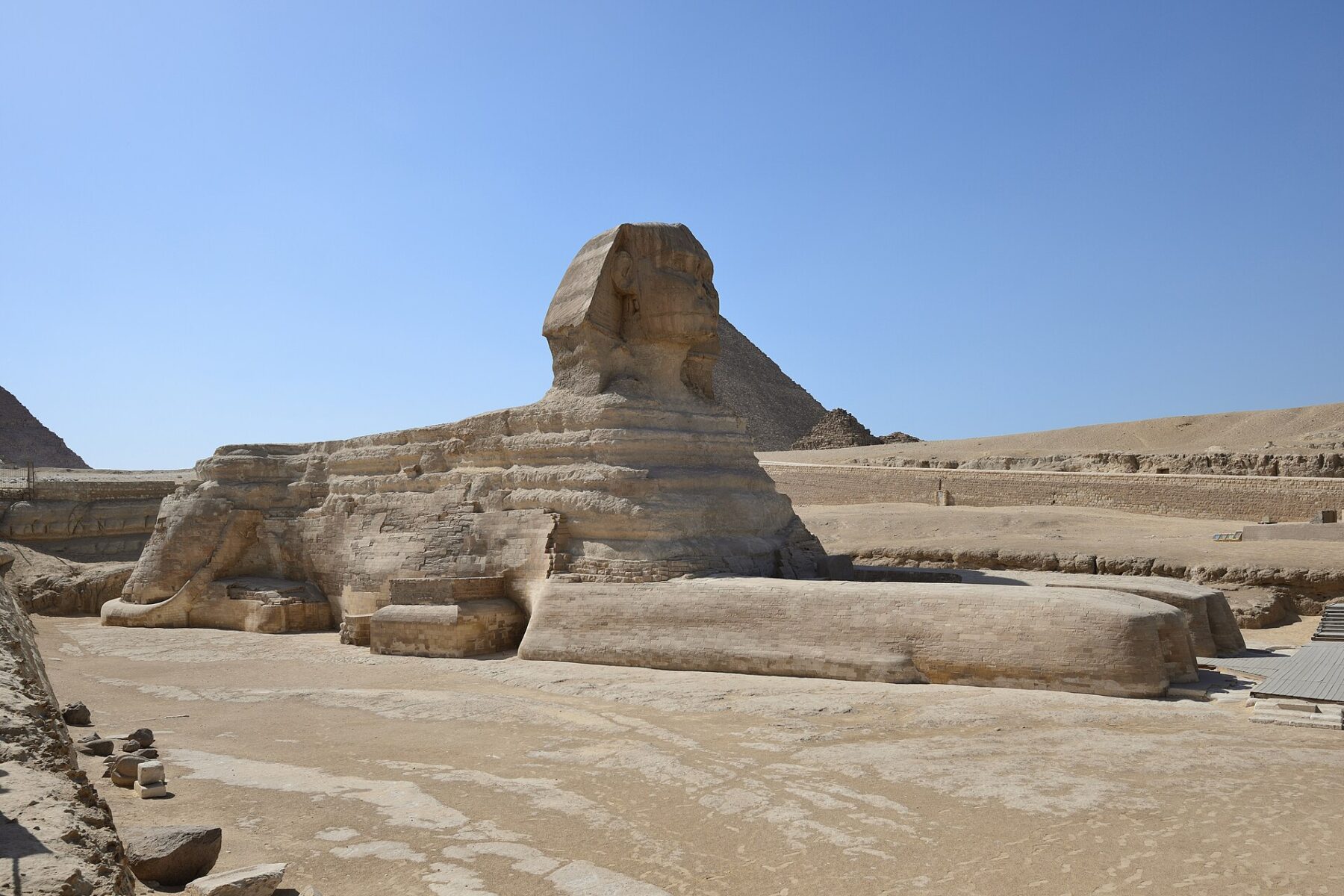 The Great Sphinx of Giza seen from the side on bare sand under a clear blue sky, with a pyramid and the plateau wall behind it