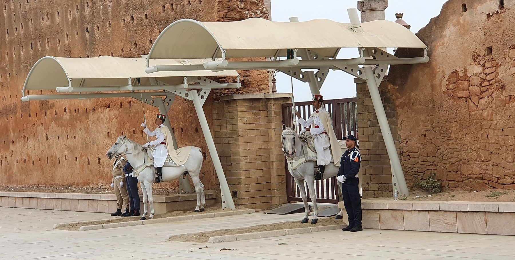 Two mounted royal guards in white uniforms sit on grey horses under pale canopies beside a red city wall, with a uniformed police officer in dark blue standing at right