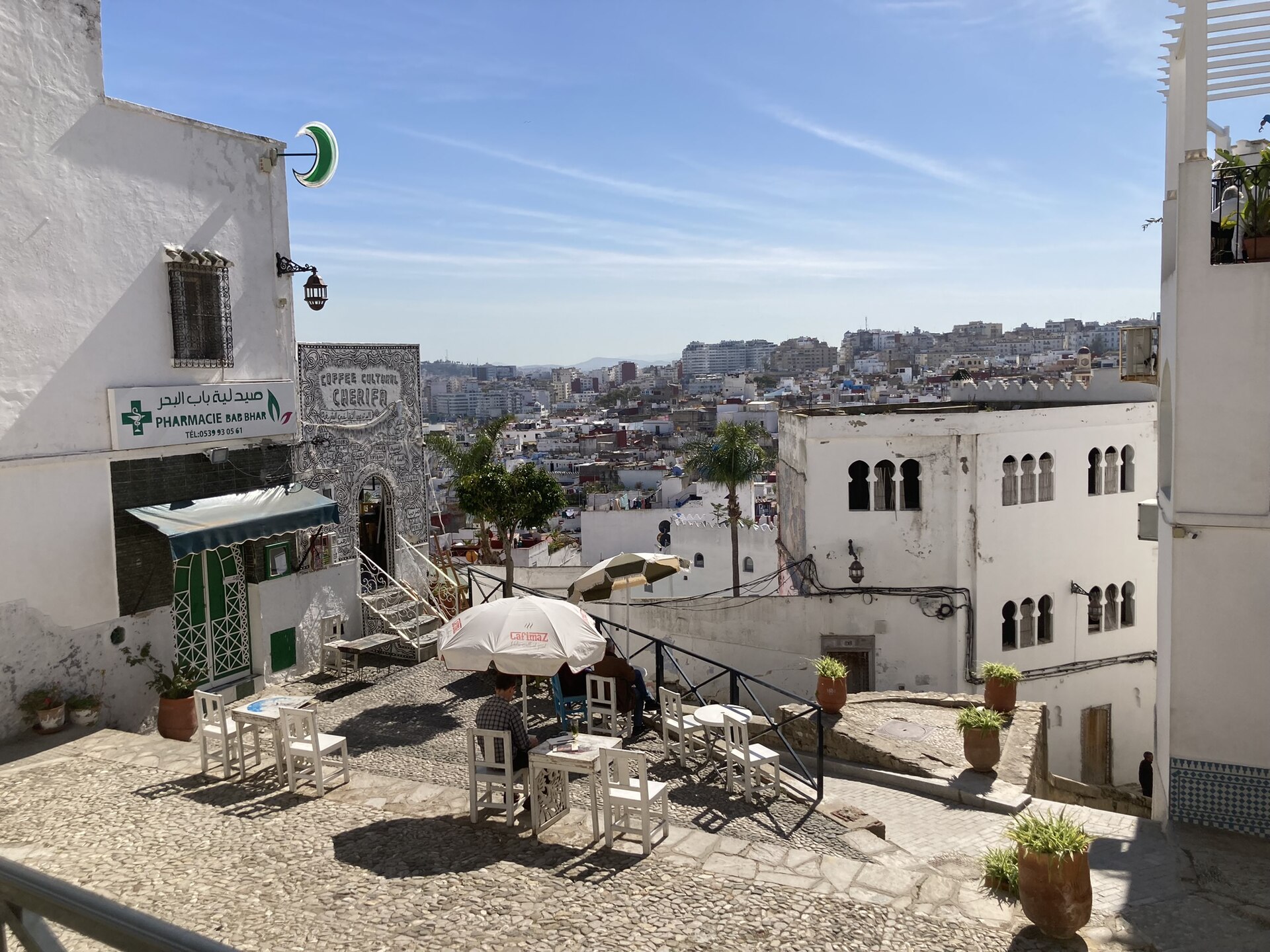 White-walled houses and a small cobbled terrace with cafe tables and parasols above the medina of Tangier, with the modern city on the hills behind