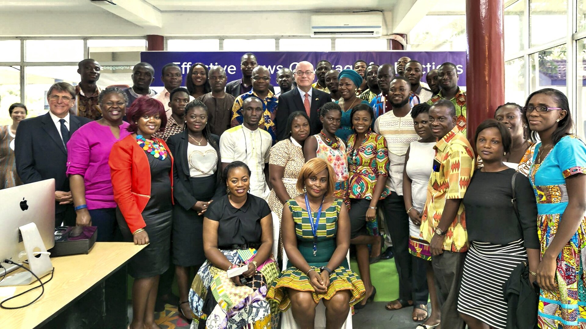 A large group of men and women, many in colourful African print clothing, posing together in a room with a banner behind them