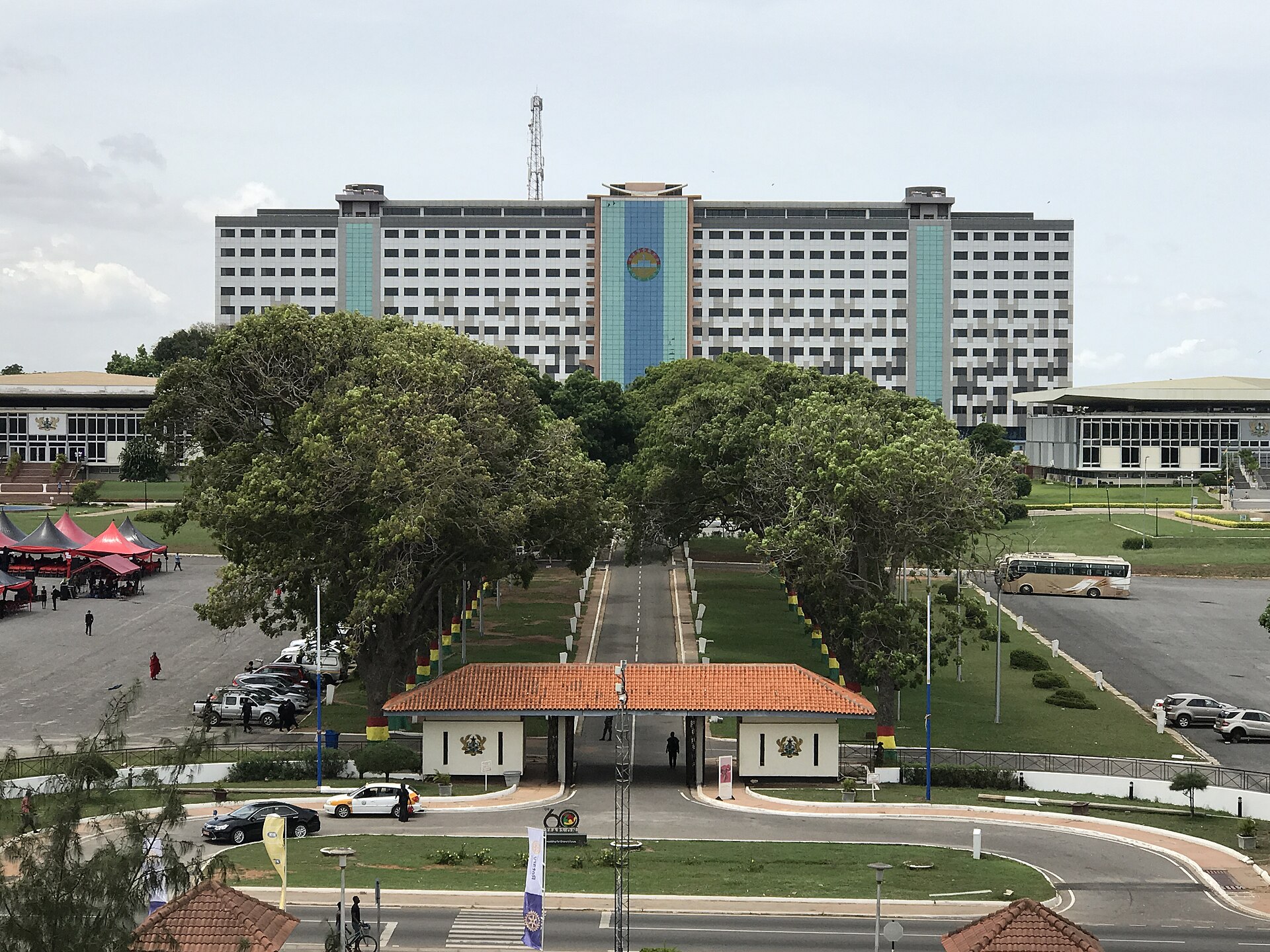 A driveway and gatehouse with an orange tiled roof leading towards a large multi-storey office building with a blue emblem panel