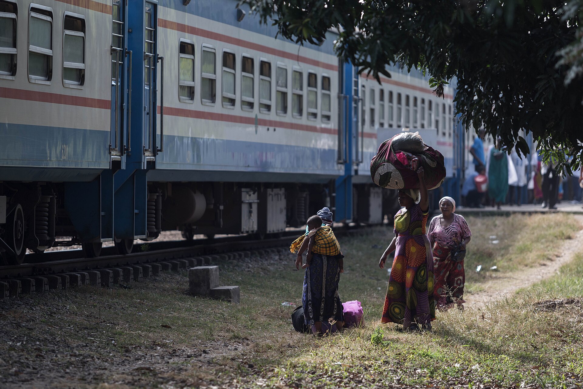 A blue and white TAZARA passenger train stands on the track while women in colourful dresses, one carrying a large bundle on her head, walk beside it in the shade of a tree