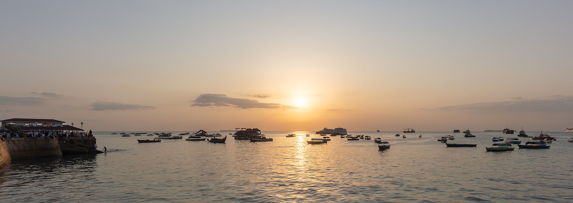 Small boats and a ferry on calm water in the harbour of Stone Town at sunset, with a quay and low buildings on the left