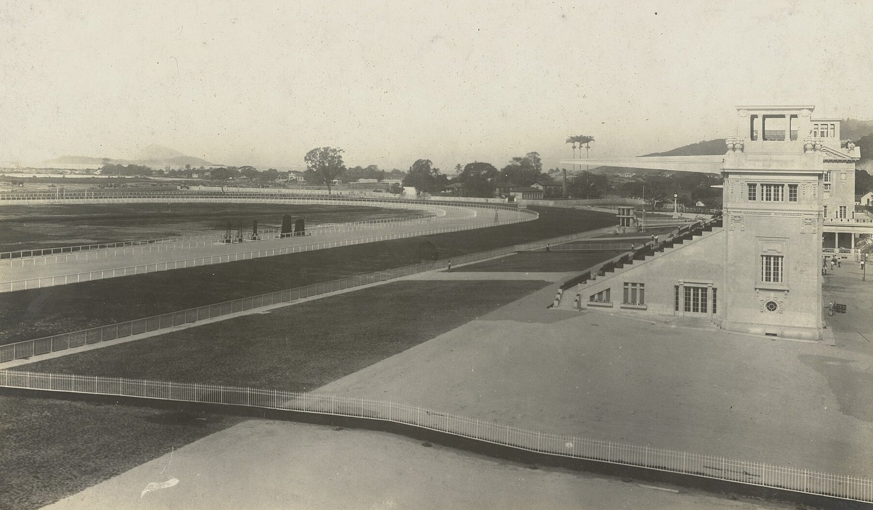 A historic black-and-white photograph of the Jockey Club racecourse in Rio de Janeiro, with the oval dirt track, a white fence and a stone grandstand tower on the right