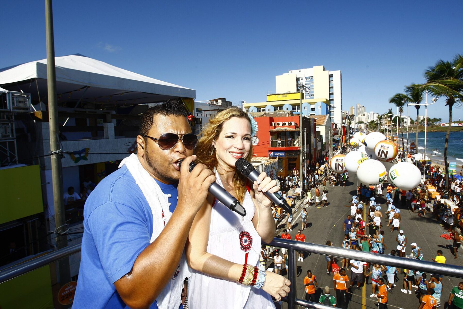 A man in sunglasses and a woman in white sing into microphones on a raised stage above a crowd on a seafront street in Salvador, with inflatable balloons, palm trees and the sea behind