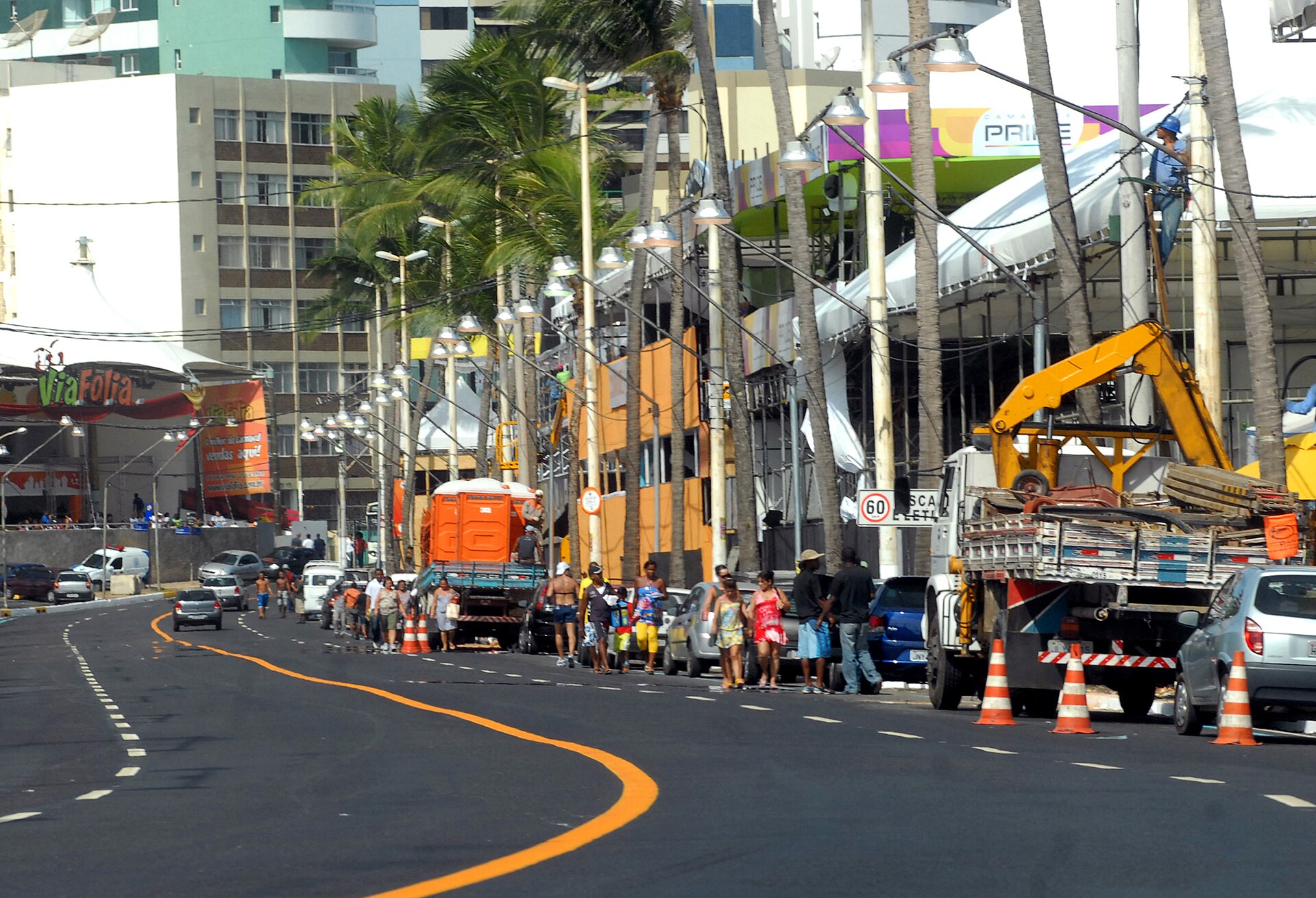 A road with an orange centre line beside tents and scaffolding under construction, workers on a lifting platform, a truck with a crane, portable toilets, palm trees and apartment blocks