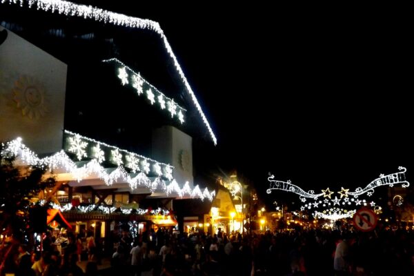 Christmas lights outline a large chalet-style building at night while a crowd fills the street, with more light arches over the road at right