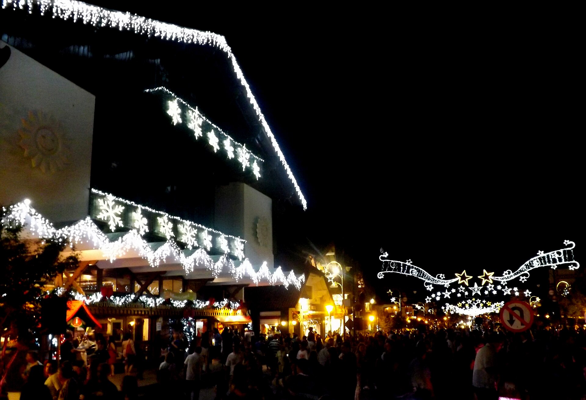 Christmas lights outline a large chalet-style building at night while a crowd fills the street, with more light arches over the road at right