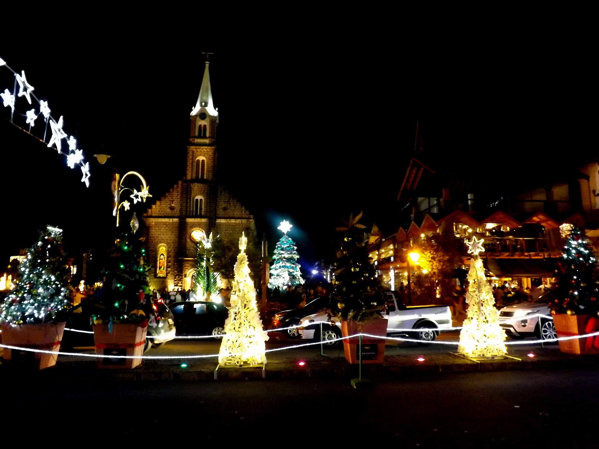 Illuminated Christmas trees in planters stand in a street at night in front of a stone church with a tall spire, with star lights at upper left