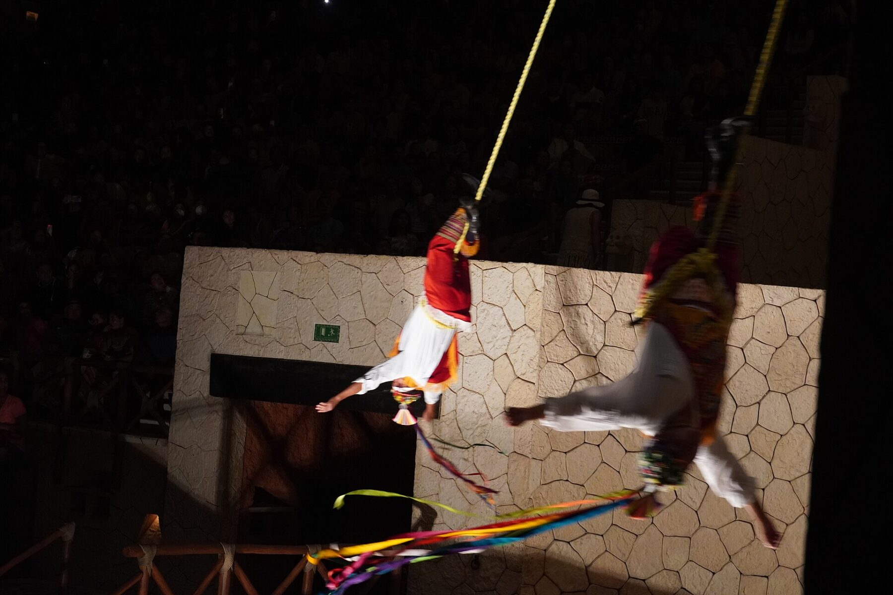 Two performers in white and red costumes hang upside down from ropes against a stone wall at an evening show at Xcaret Park, with a dark audience behind them