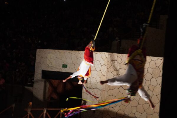 Two performers in white and red costumes hang upside down from ropes against a stone wall at an evening show at Xcaret Park, with a dark audience behind them