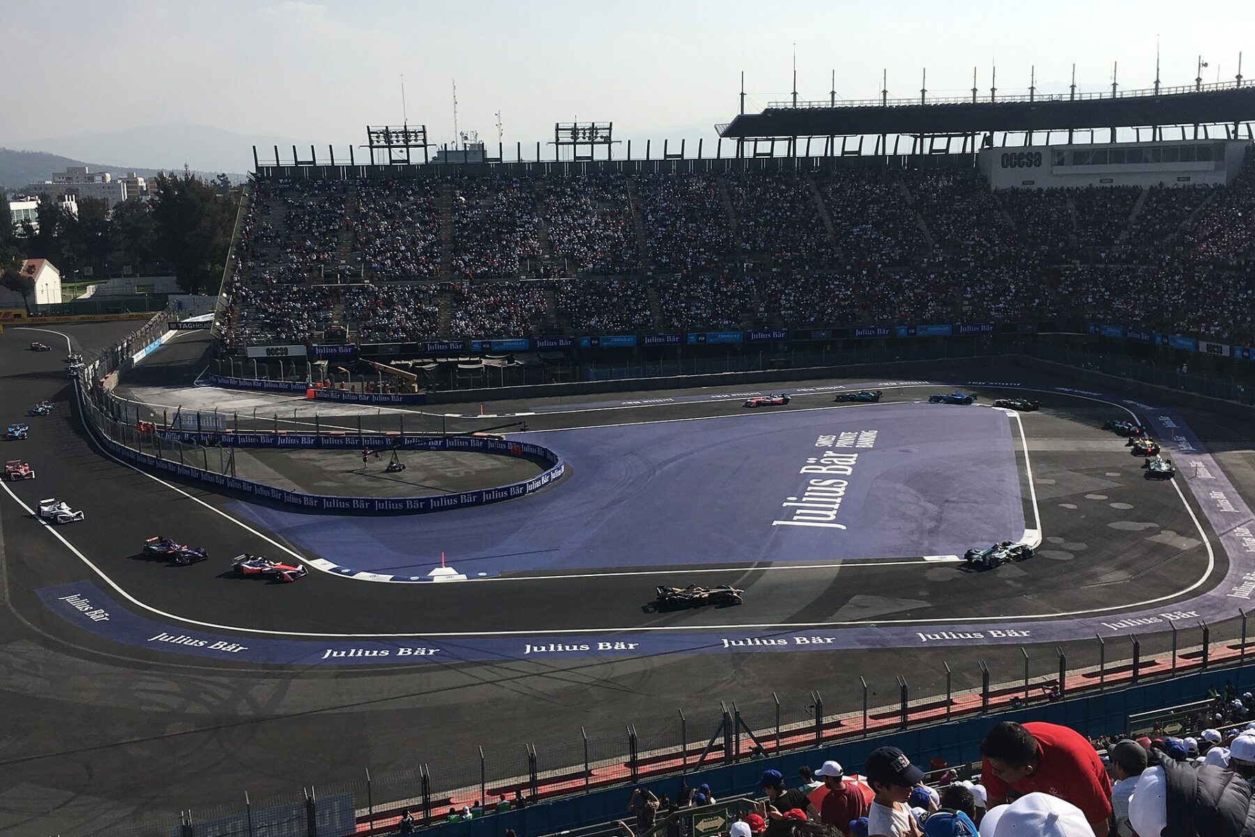 Formula E race cars rounding a curve on a blue-branded track inside a packed stadium section, with spectators in the foreground