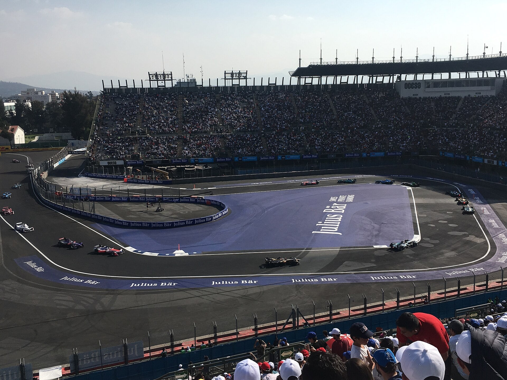 Formula E race cars rounding a curve on a blue-branded track inside a packed stadium section, with spectators in the foreground