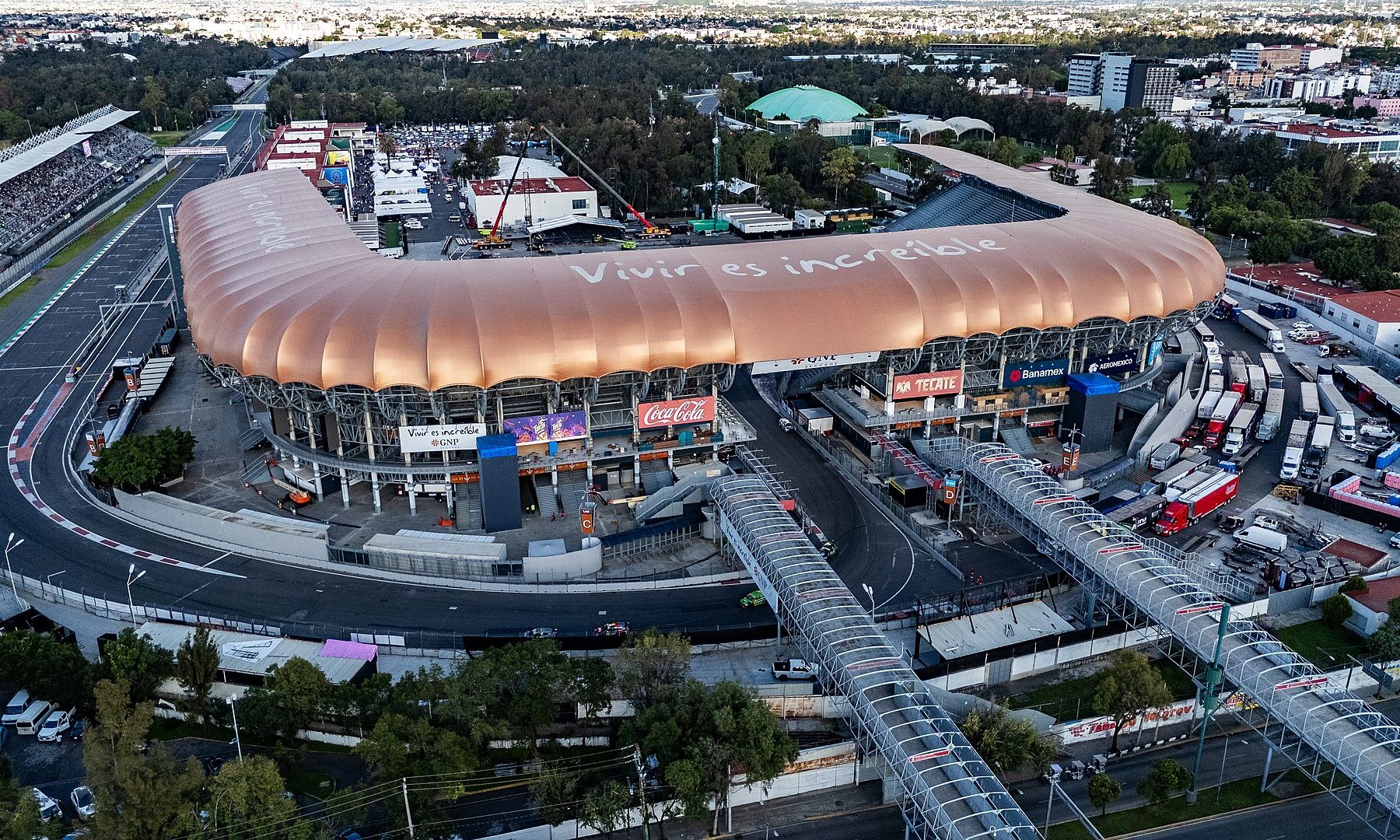 Drone view of Estadio GNP Seguros in Mexico City, a stadium with a curved copper-coloured roof reading Vivir es increíble, ringed by the Autódromo Hermanos Rodríguez racetrack and footbridges