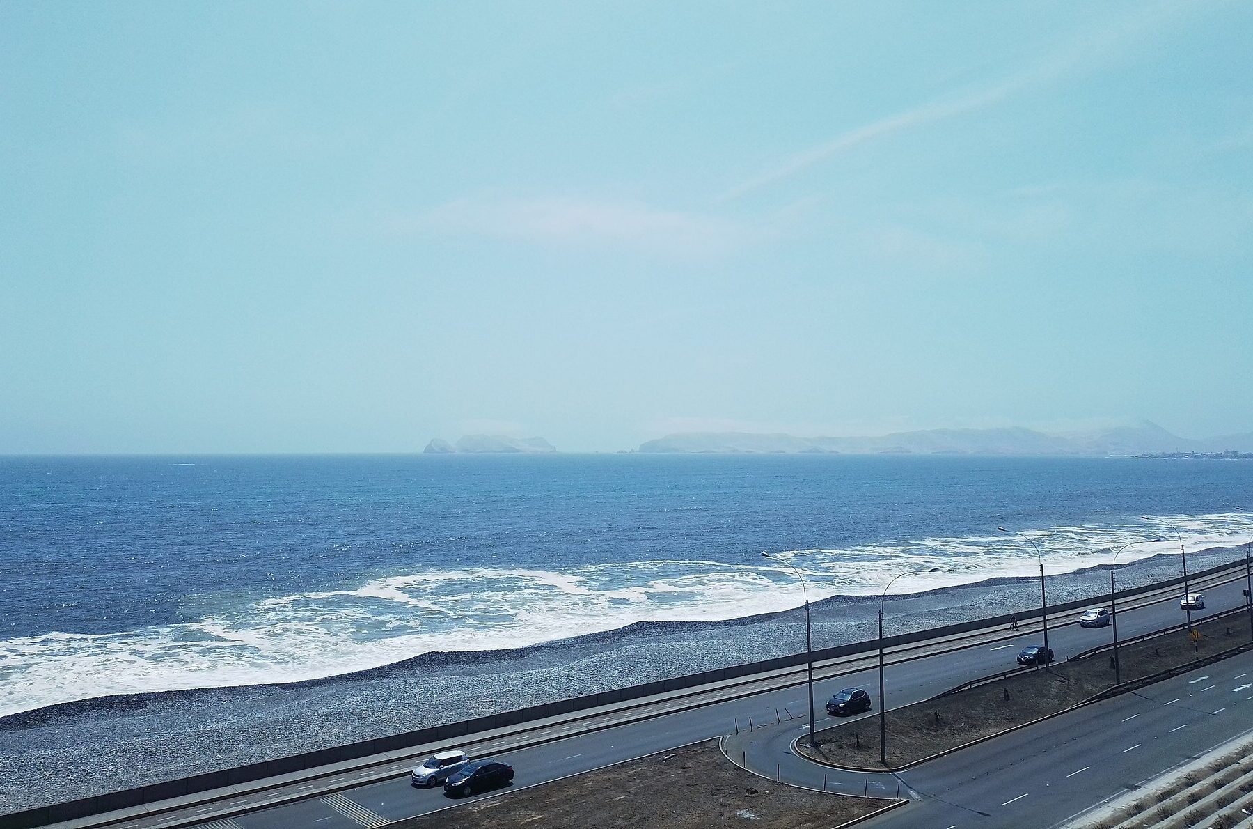 A coastal highway with a few cars runs beside a pebble beach and breaking waves, with islands on the horizon under a pale sky
