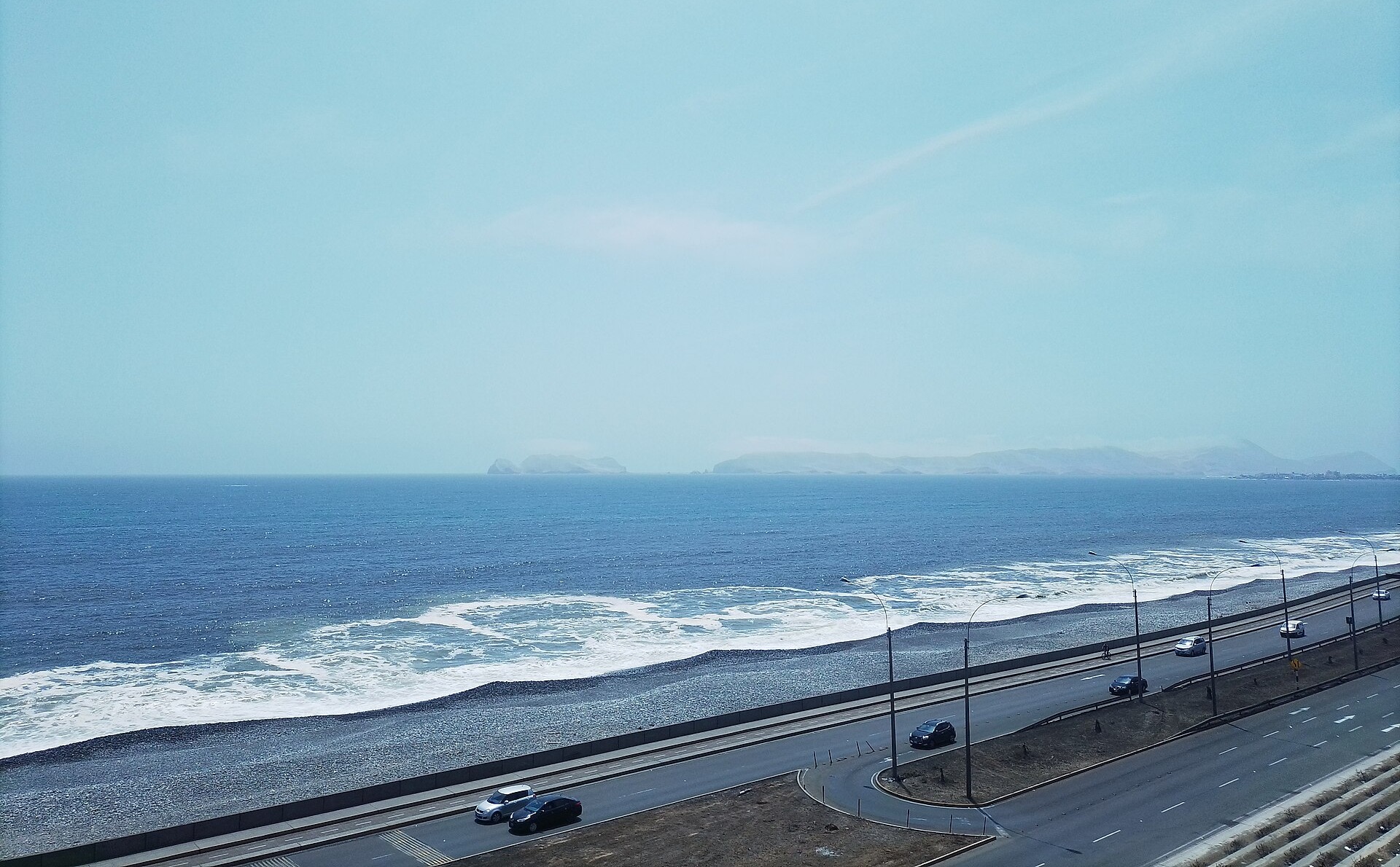 A coastal highway with a few cars runs beside a pebble beach and breaking waves, with islands on the horizon under a pale sky