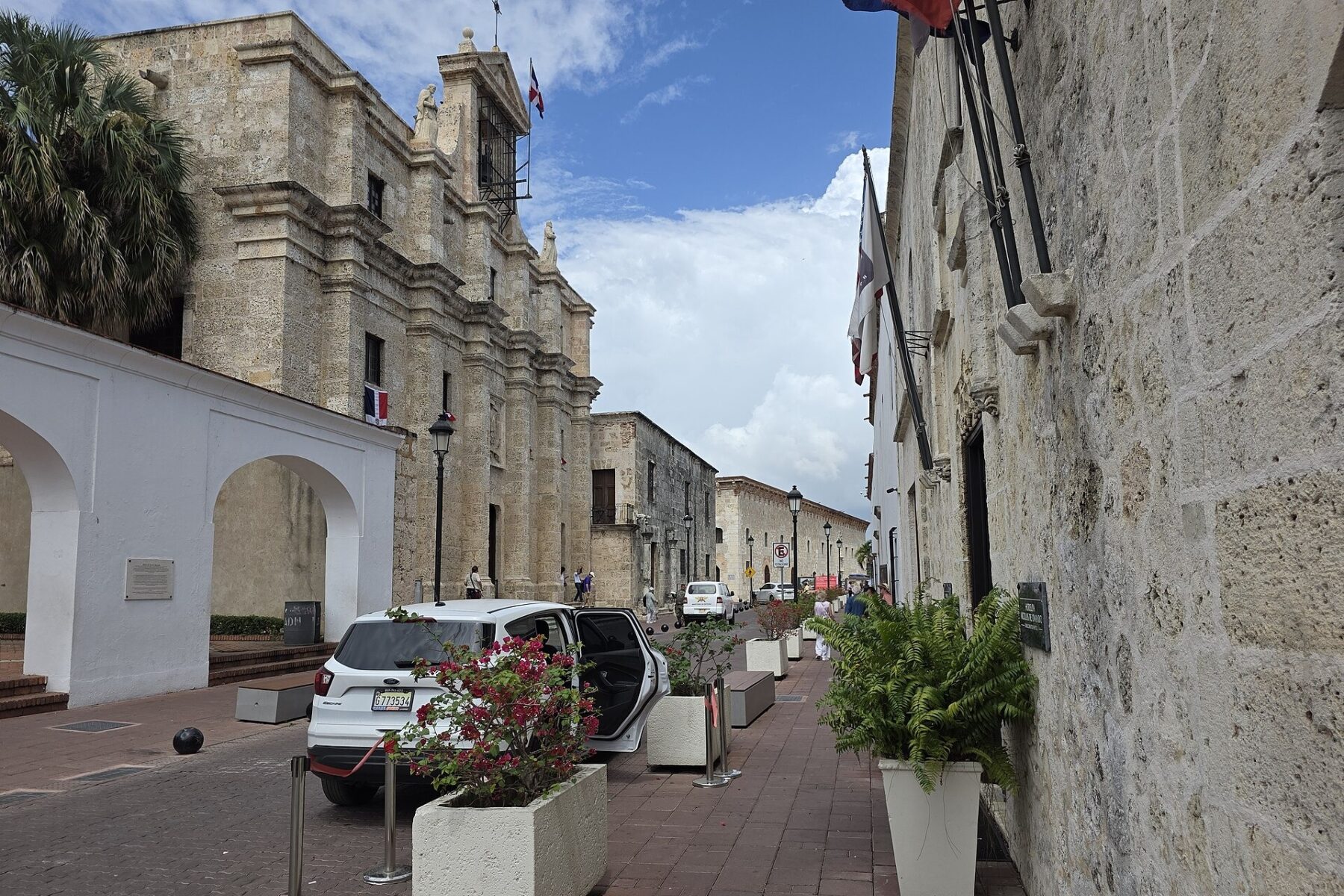 Stone colonial buildings with flags and planters line a brick-paved street under a blue sky, with a parked white car