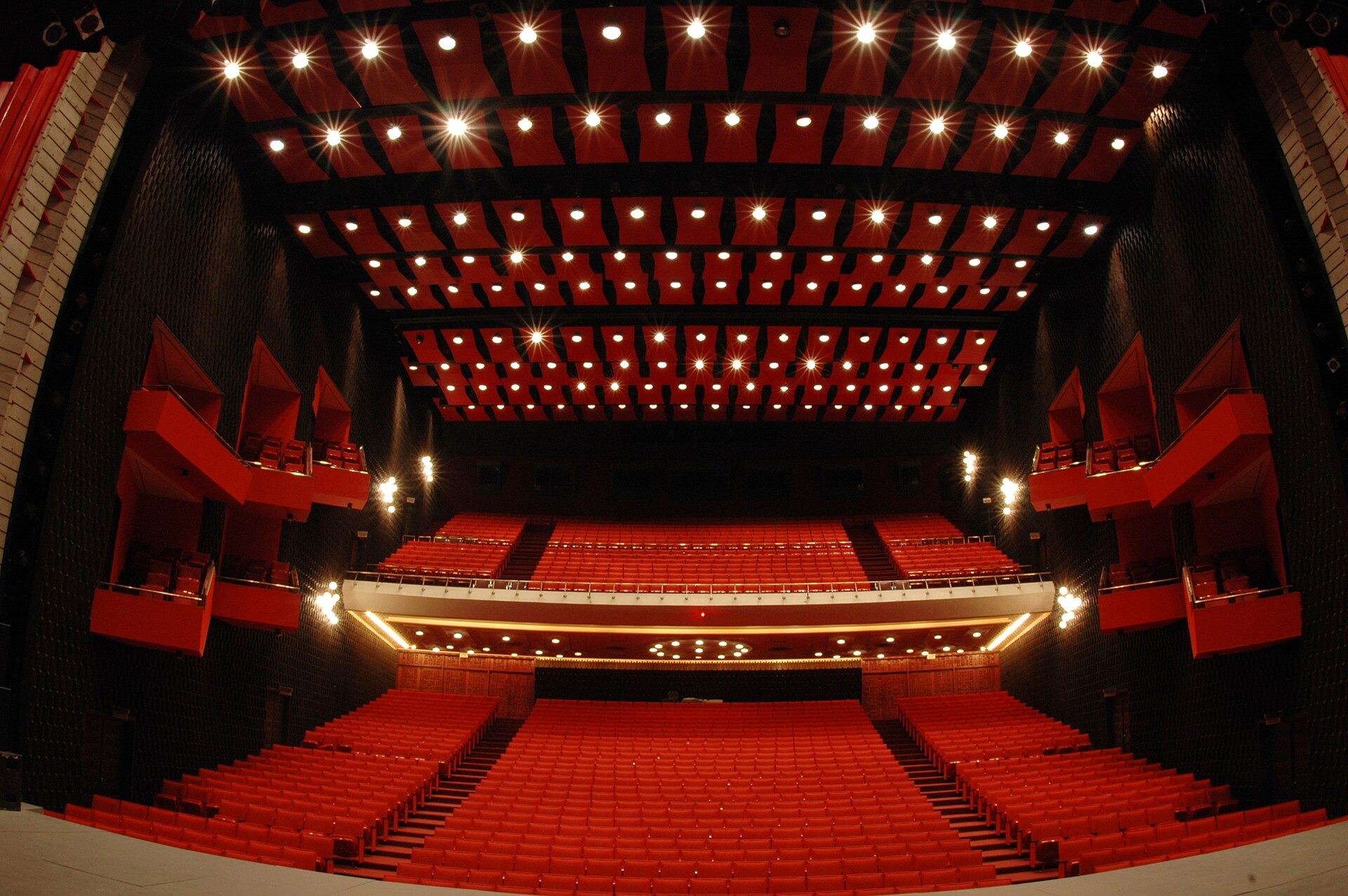 A large theatre hall with rows of red seats, a curved balcony and red side boxes under a ceiling of red panels set with small lights
