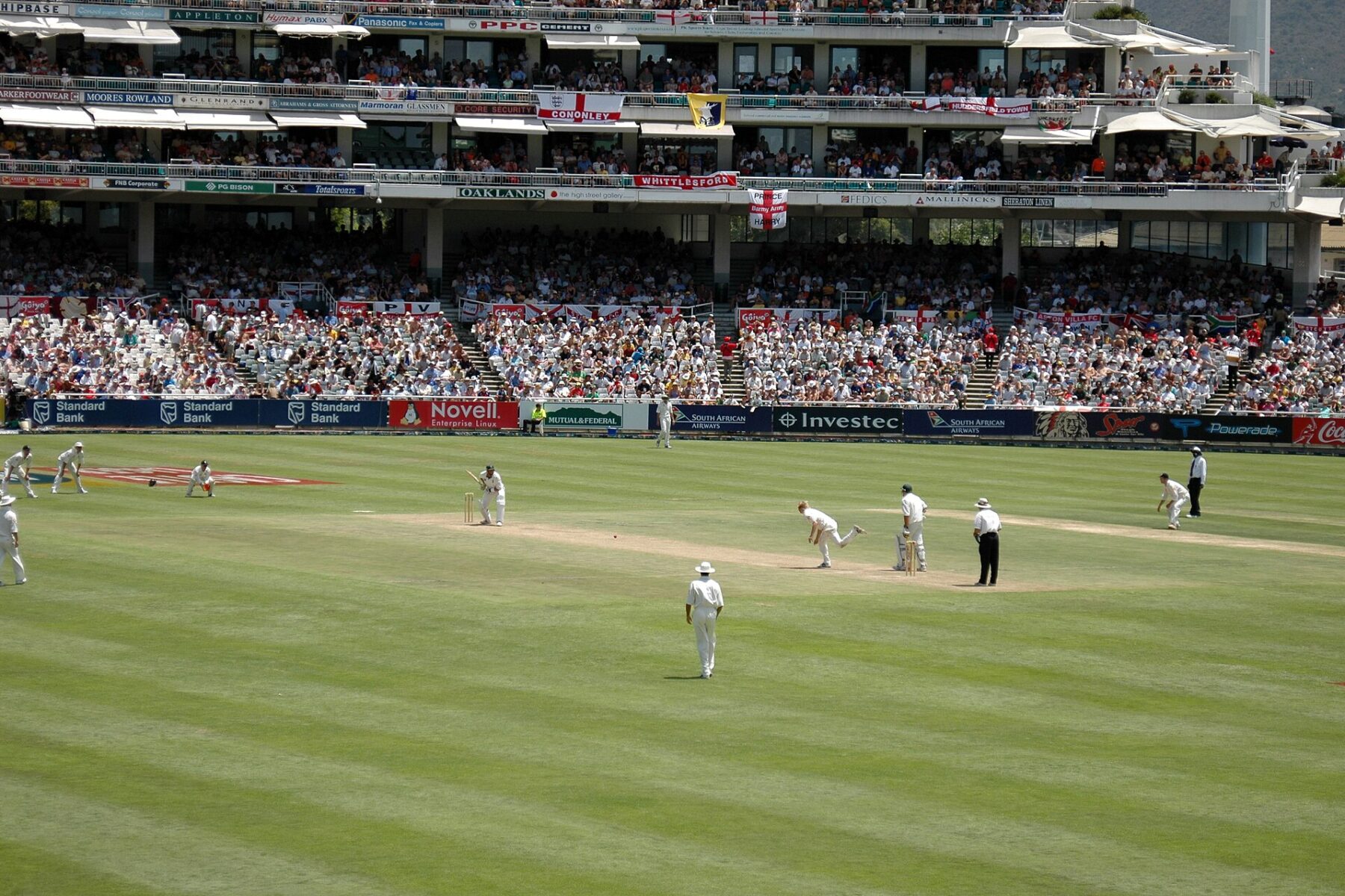 A Test match in progress at Newlands in Cape Town, with a bowler delivering to a batter, fielders in white on a green outfield and full stands hung with England flags