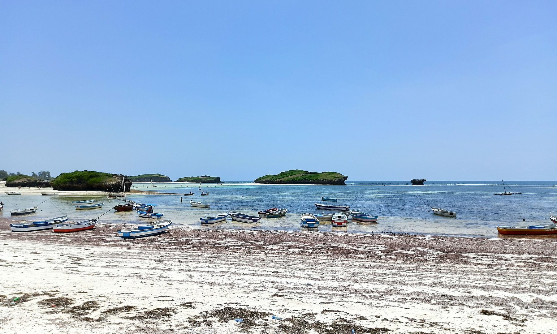 Small fishing boats rest on white sand at low tide in a shallow bay, with green rock islets and a calm blue sea beyond under a clear sky