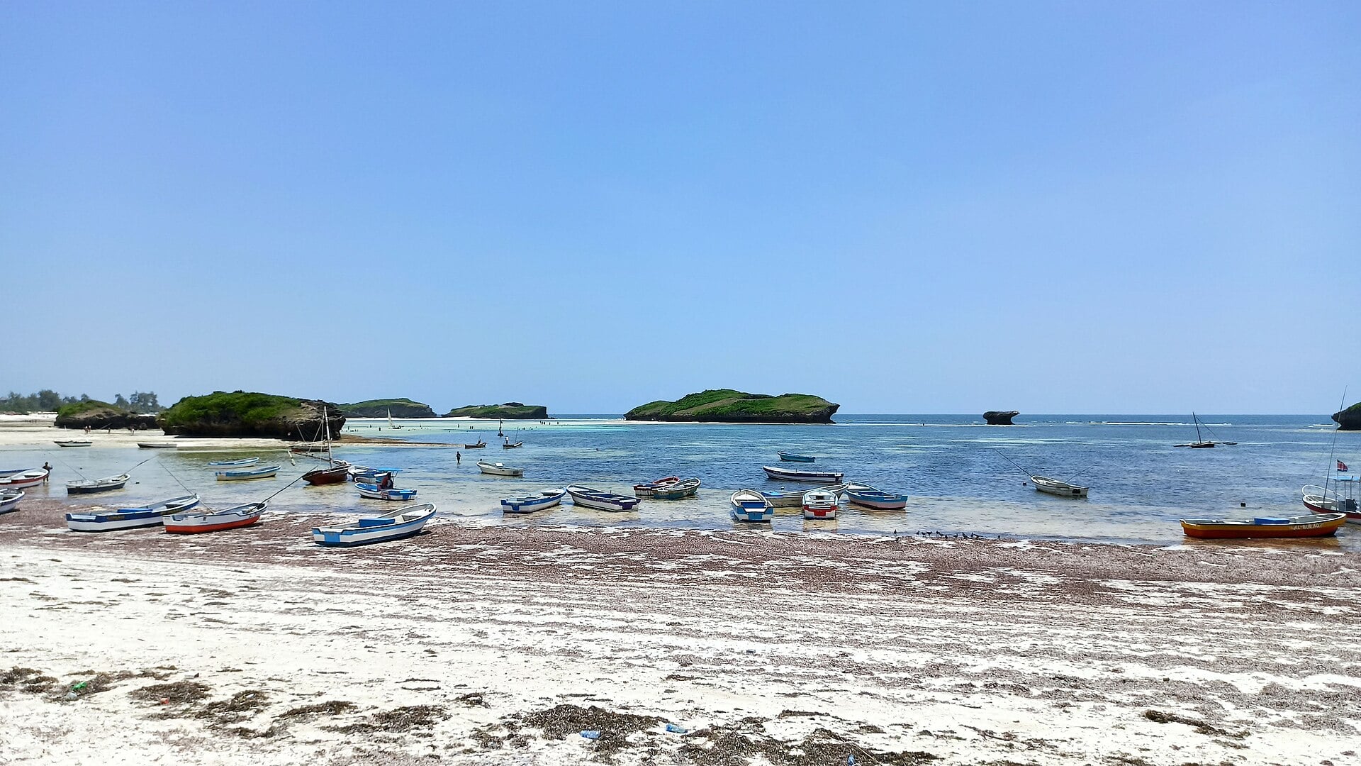 Small fishing boats rest on white sand at low tide in a shallow bay, with green rock islets and a calm blue sea beyond under a clear sky