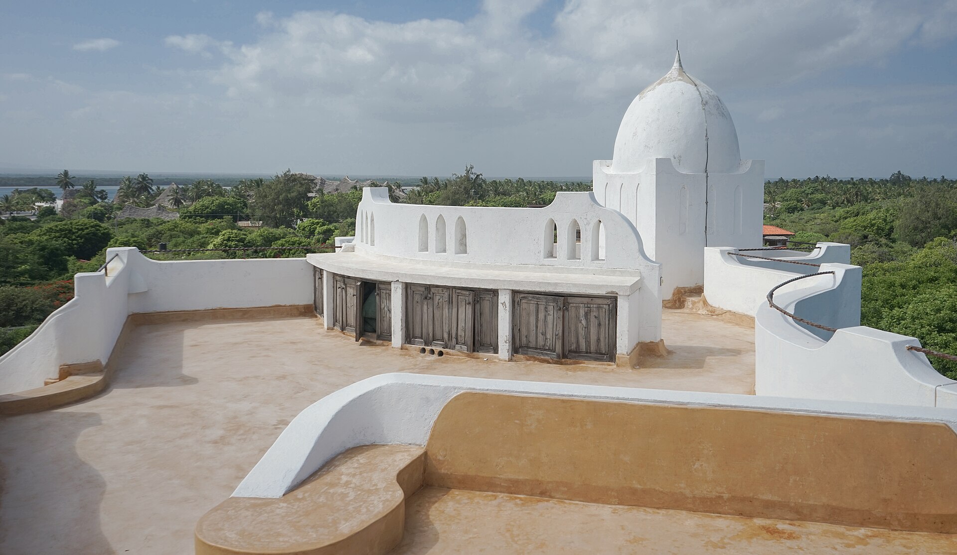 A white domed building with a flat rooftop terrace, surrounded by green trees, with the sea on the horizon