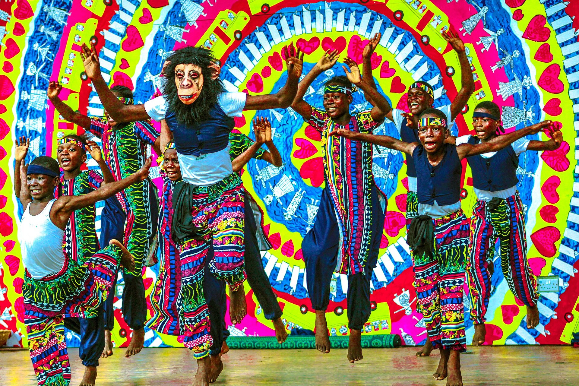 Young dancers in headbands and brightly patterned costumes, one wearing an ape mask, leap on a stage in front of a colourful heart-patterned backdrop at a festival in Freedom Park, Lagos