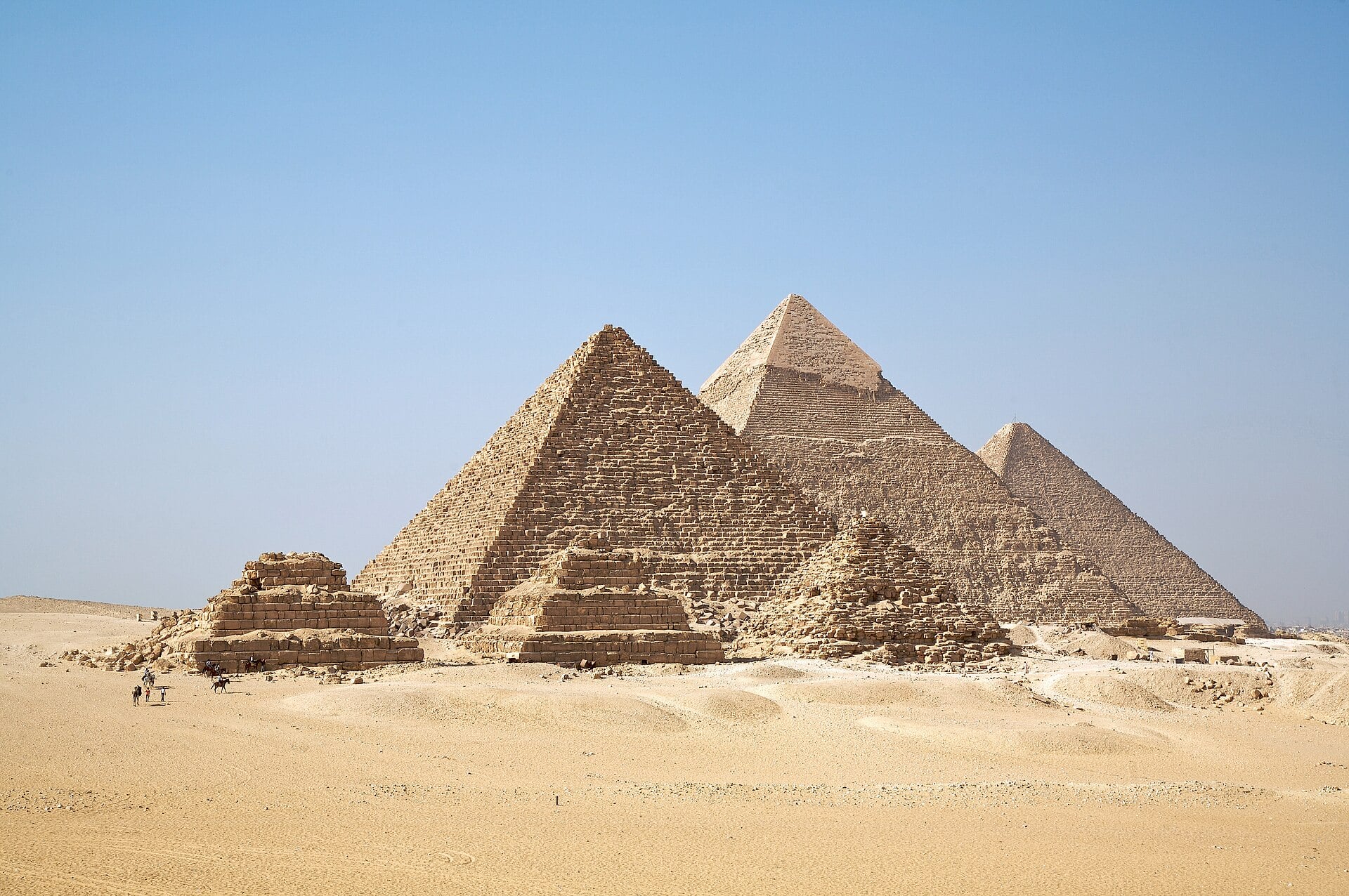 The three large Giza pyramids and three smaller queens' pyramids rise from golden desert sand under a clear blue sky, with a few small riders and walkers in the foreground