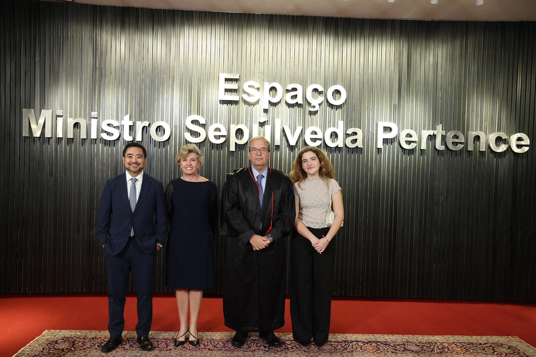 A man in a black judicial robe stands with three people in front of a dark wall with the words Espaço Ministro Sepúlveda Pertence
