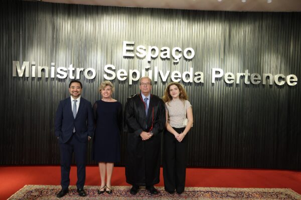 A man in a black judicial robe stands with three people in front of a dark wall with the words Espaço Ministro Sepúlveda Pertence