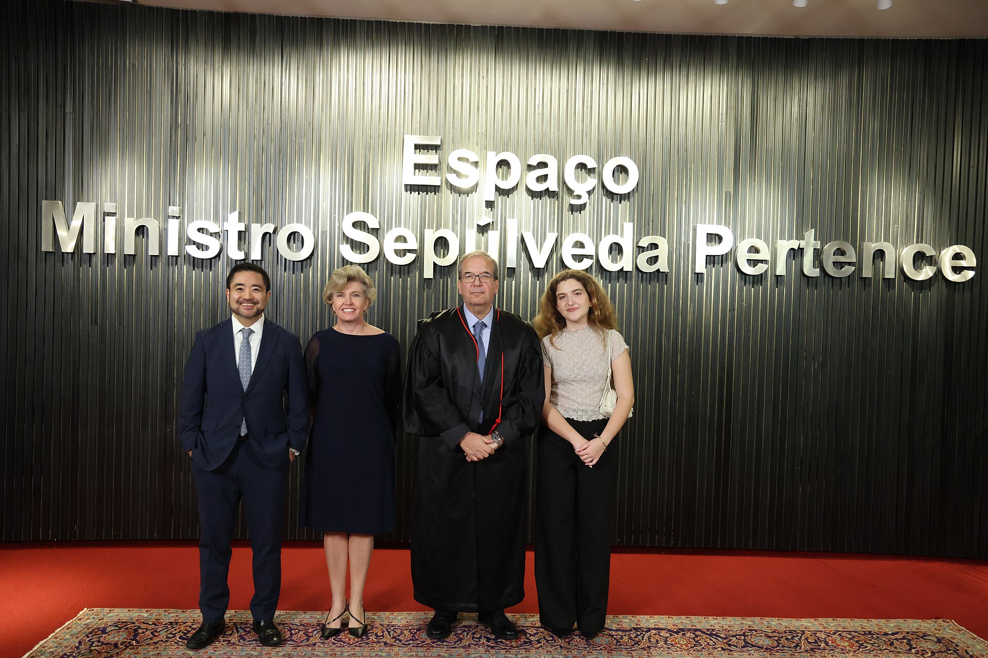 A man in a black judicial robe stands with three people in front of a dark wall with the words Espaço Ministro Sepúlveda Pertence