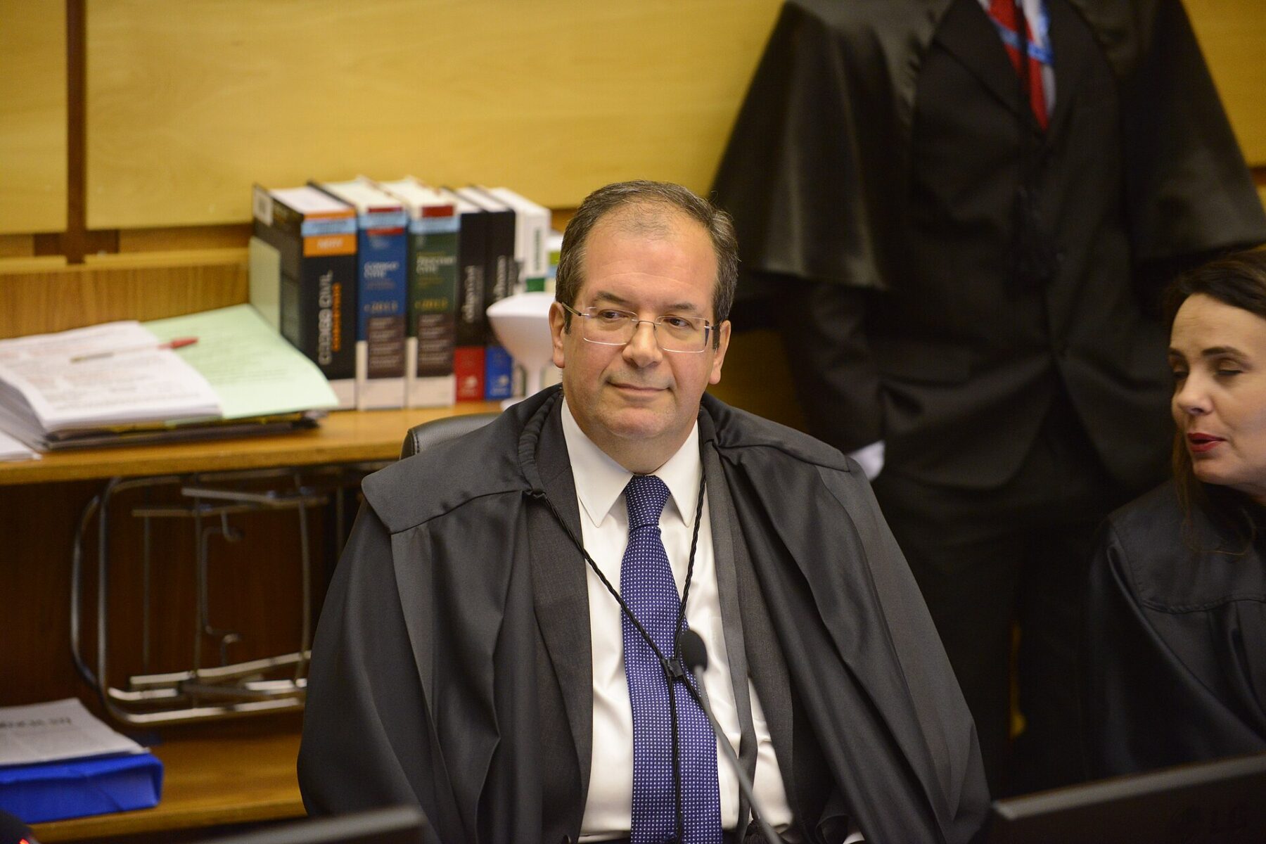 A man with glasses in a black judicial robe and blue tie sits at a bench in a wood-panelled courtroom with law books behind him