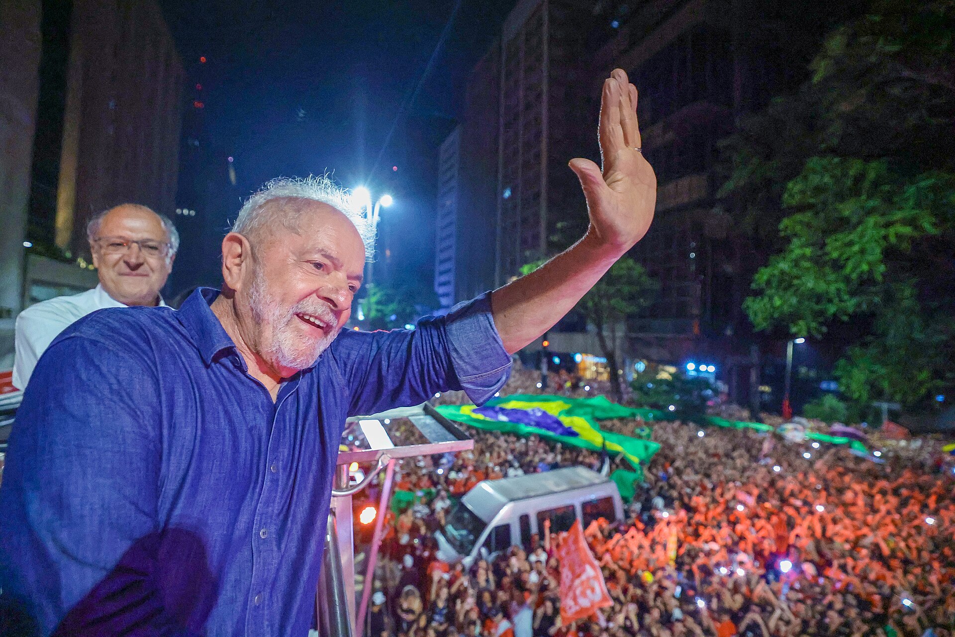 A smiling grey-haired man in a blue shirt waves from a stage at night, with a bespectacled man behind him and a large crowd with a Brazilian flag below
