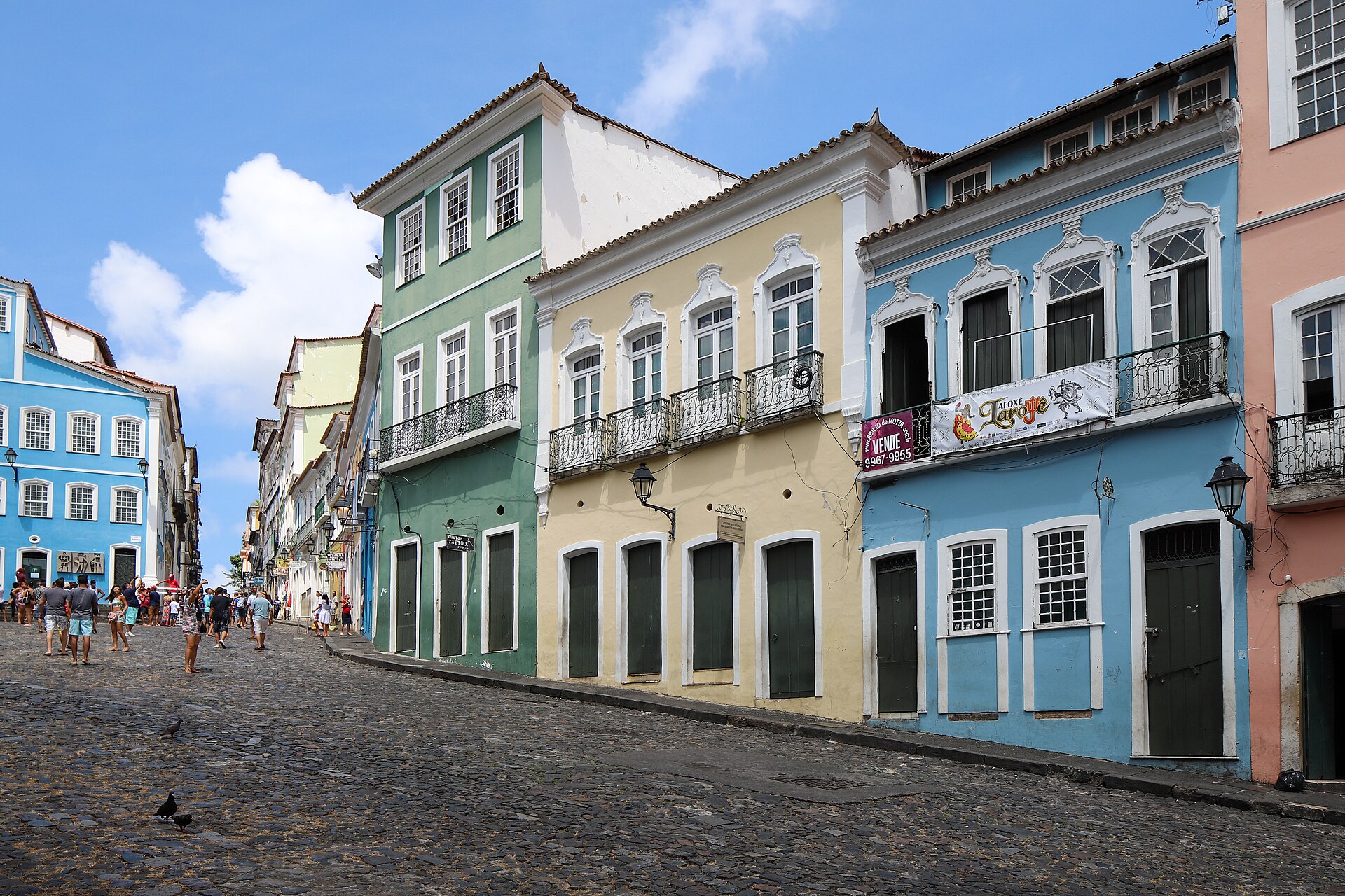 Colourful two- and three-storey colonial houses with balconies line a sloping cobbled square, with visitors walking in the distance