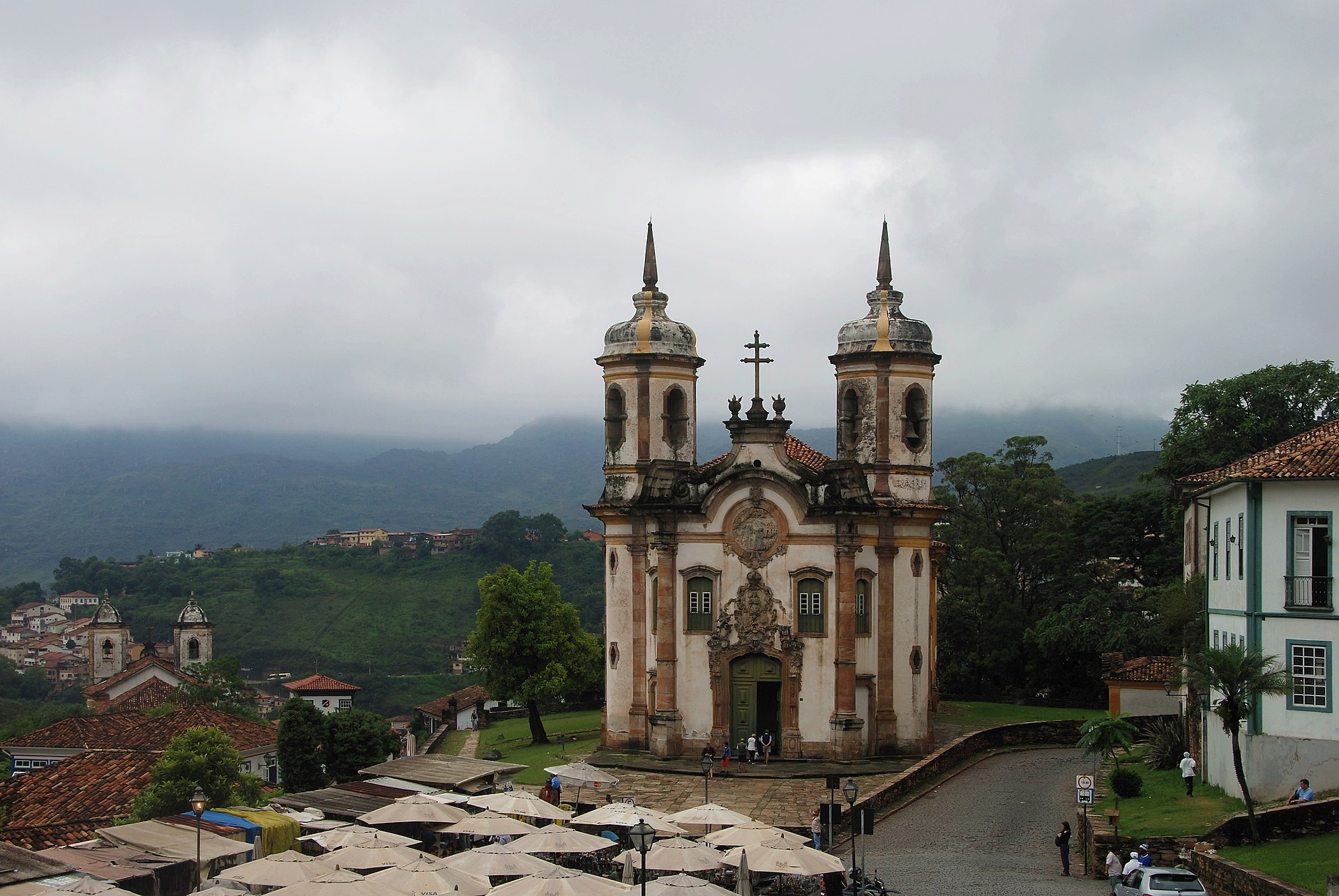 A white baroque church with two round bell towers stands above market umbrellas, with green hills and low clouds behind