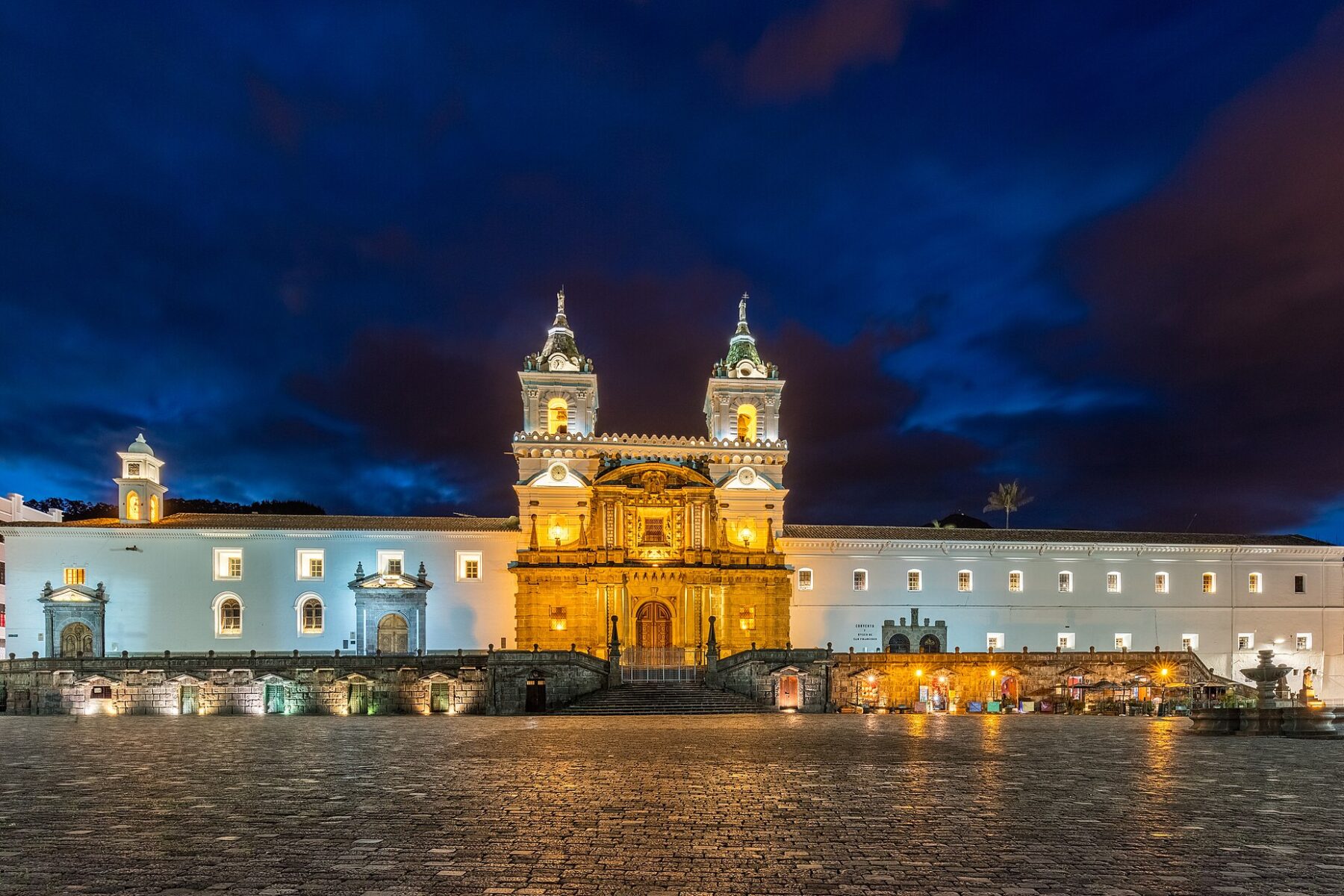 The white facade and twin bell towers of the San Francisco church and monastery in Quito, lit up at dusk above an empty plaza