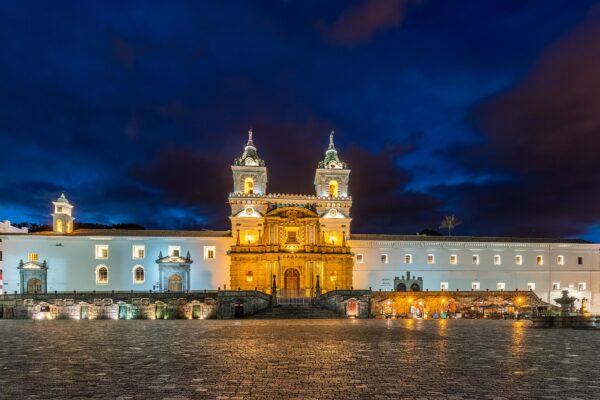 The white facade and twin bell towers of the San Francisco church and monastery in Quito, lit up at dusk above an empty plaza