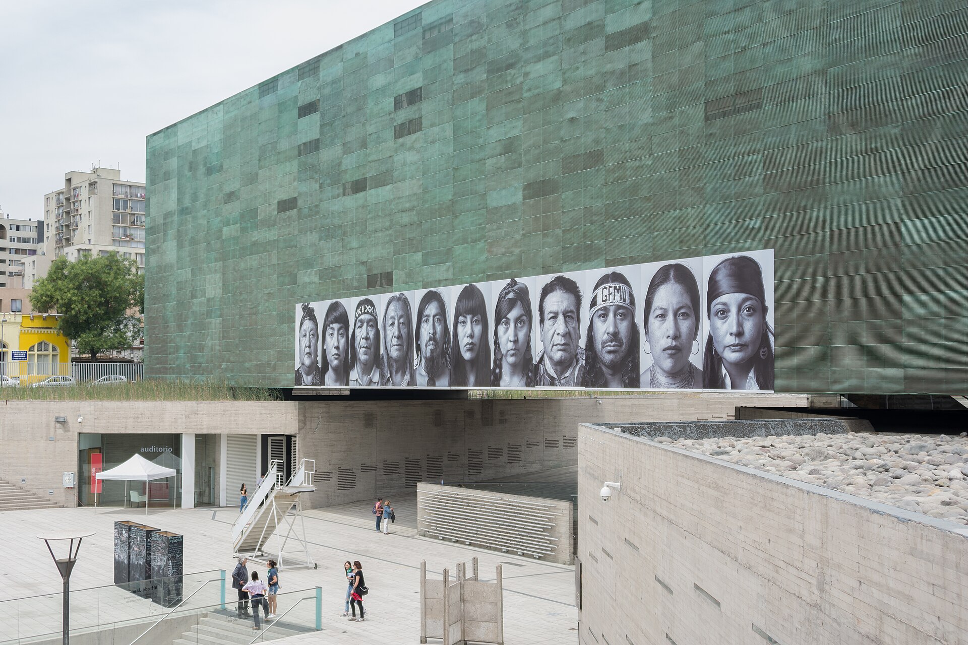 The green copper-clad facade of the Museum of Memory and Human Rights in Santiago, with a row of large portrait photographs above a concrete plaza