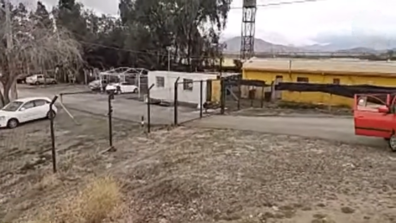 A fenced yard with parked cars and low yellow and white buildings at the entrance area of the Punta Peuco prison, with hills behind
