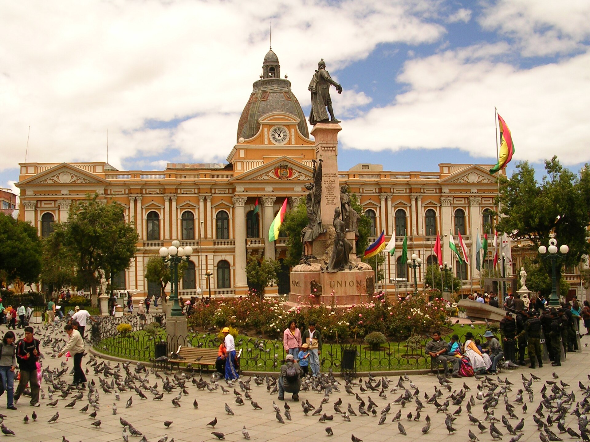 Plaza Murillo in La Paz with a stone monument, pigeons, pedestrians, flags and a yellow government building with a clock dome