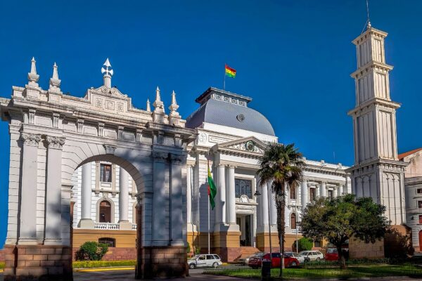 The white neoclassical Supreme Court building in Sucre with a grey dome, a Bolivian flag, a stone arch and a clock tower under a deep blue sky