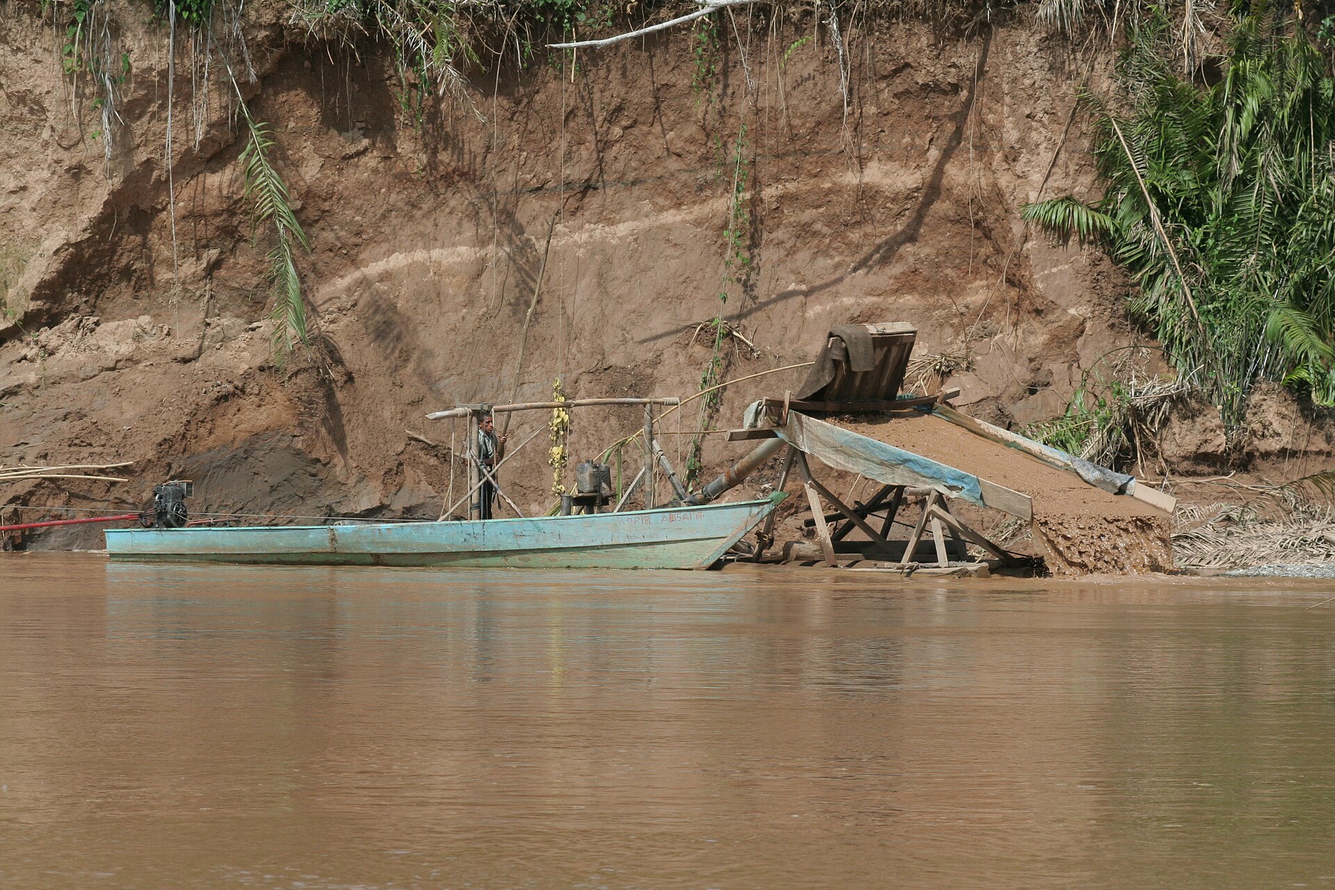Small wooden suction dredge and boat working an eroded riverbank on a brown river