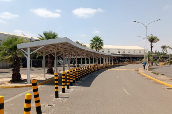 Covered walkway and bollard-lined road outside a terminal building at Jomo Kenyatta International Airport in Nairobi, with palm trees under a partly cloudy sky