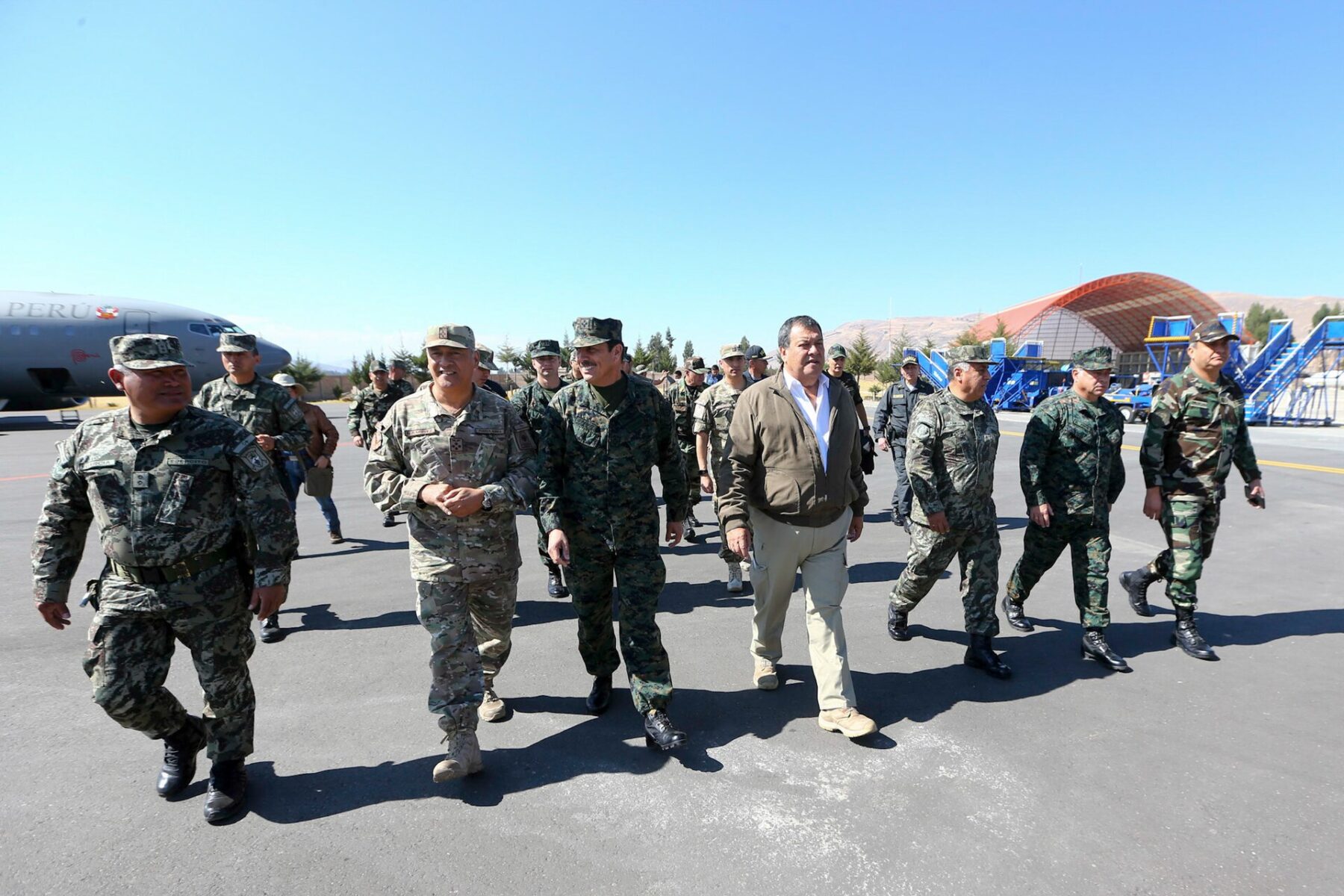 Peruvian military officers in camouflage and a civilian in a brown jacket walk across an airfield apron, with a Peruvian Air Force plane behind them