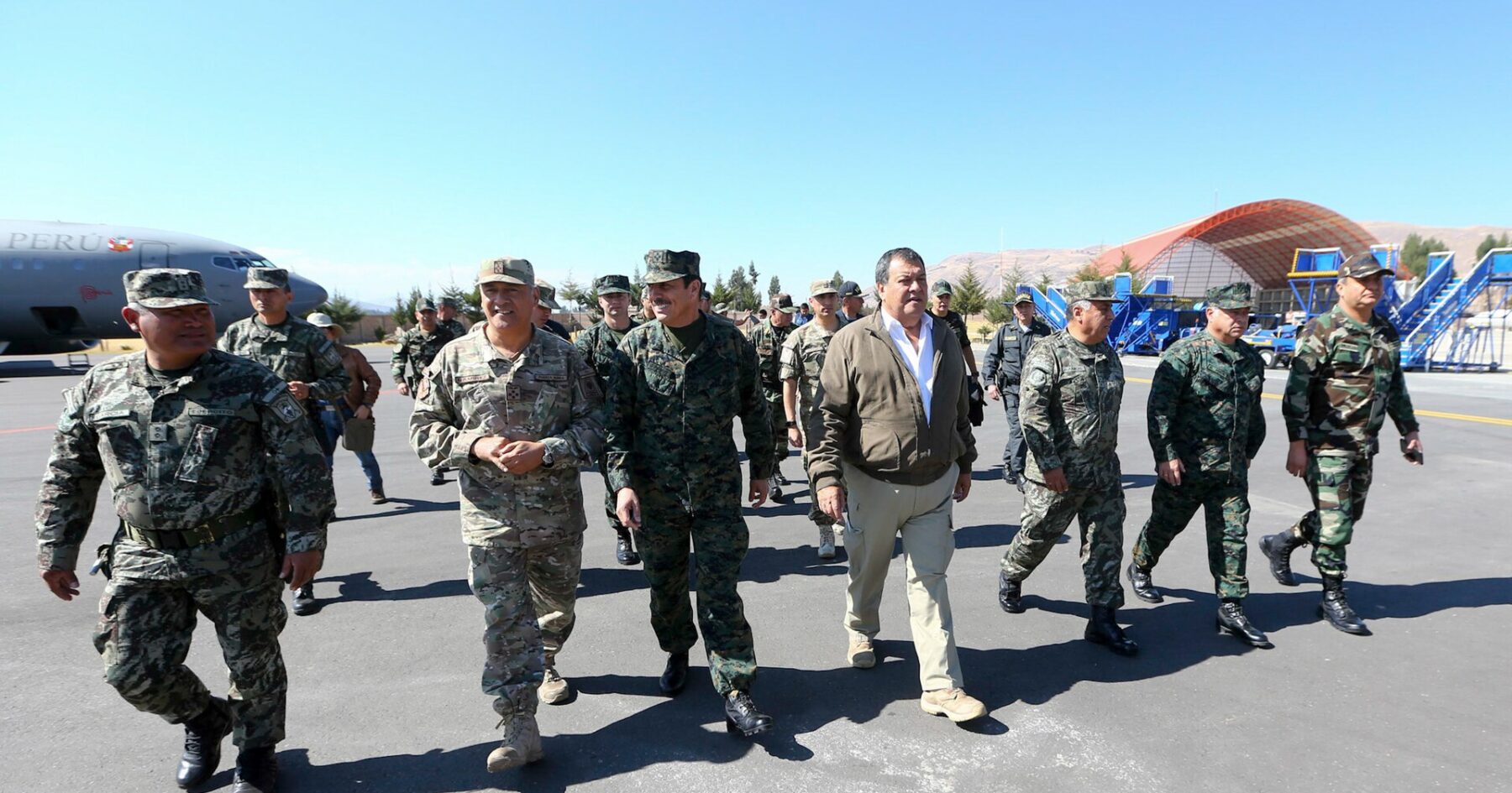 Peruvian military officers in camouflage and a civilian in a brown jacket walk across an airfield apron, with a Peruvian Air Force plane behind them