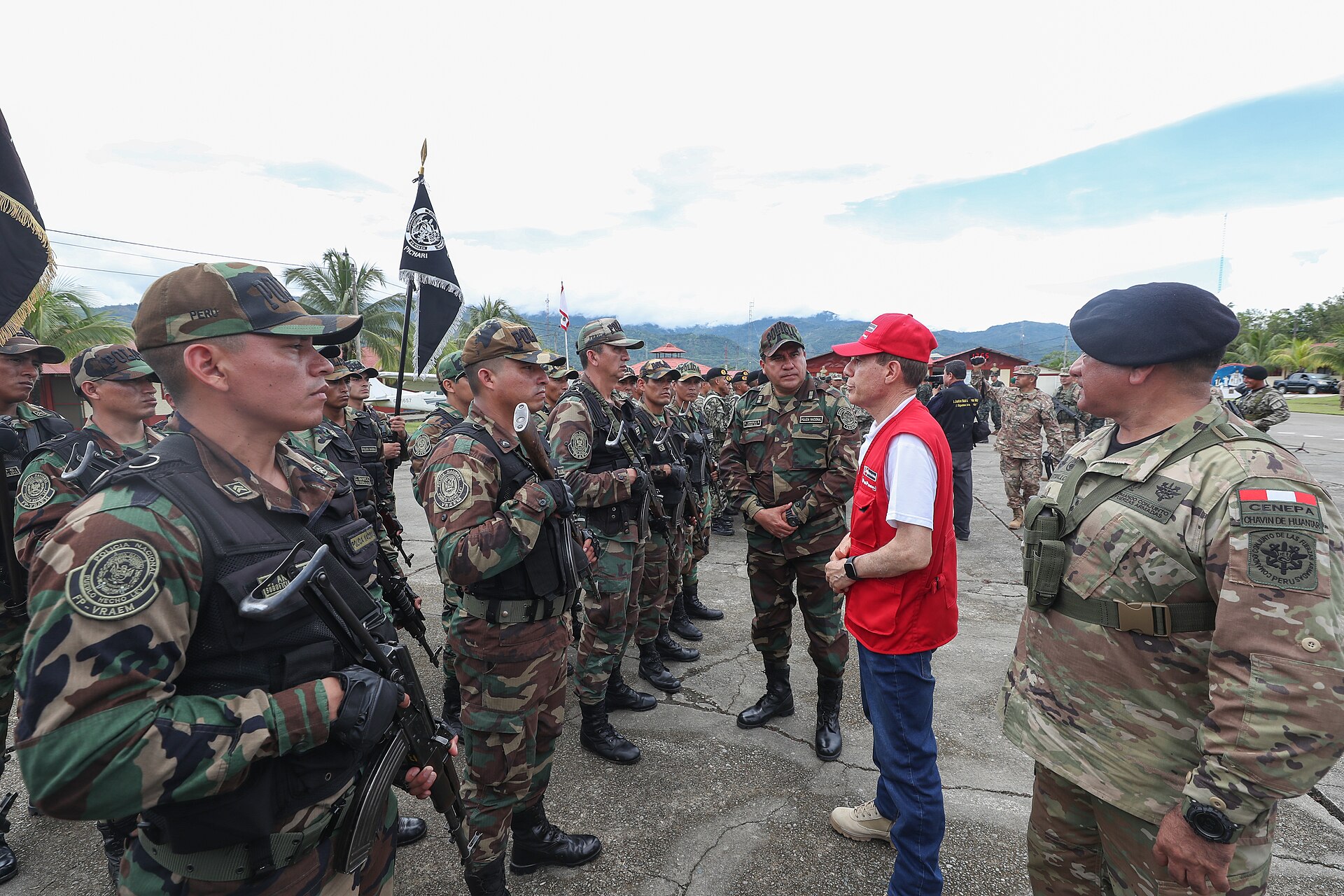 Armed Peruvian soldiers in camouflage stand in line while a man in a red vest and cap speaks with an officer