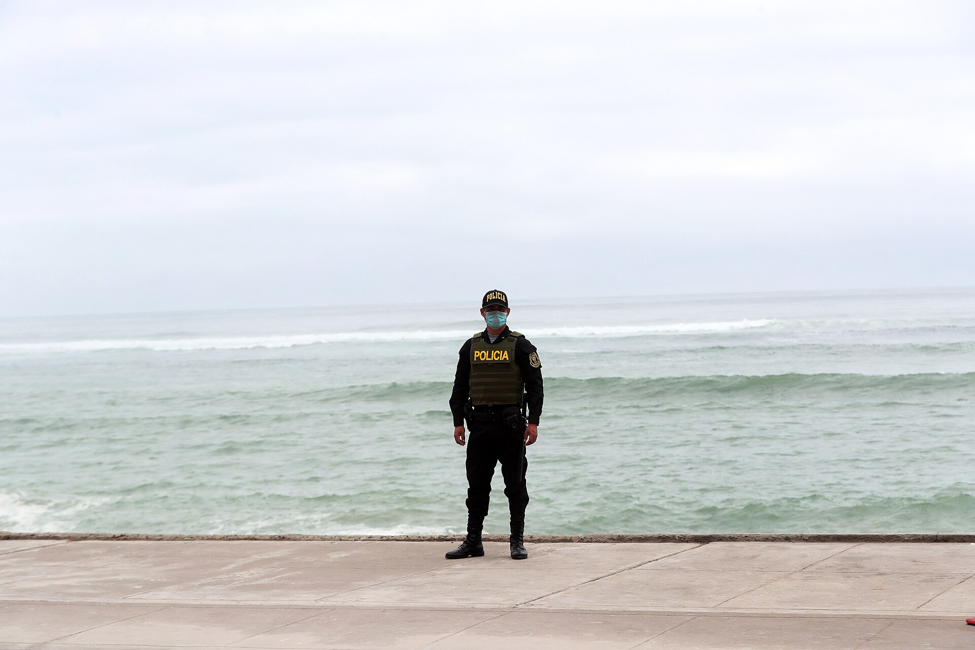 A Peruvian police officer in a vest marked Policia stands on a seafront promenade in front of the Pacific Ocean