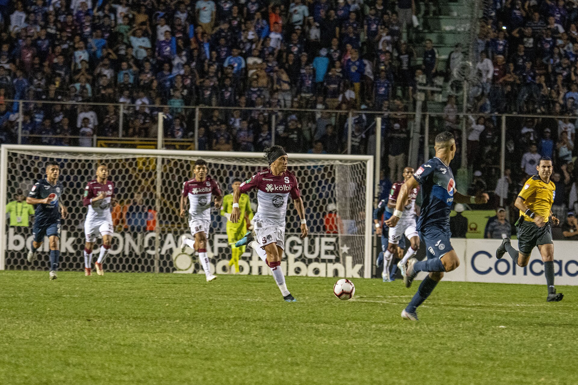 Players in blue and maroon kits chase a football in front of a goal and packed stands during a night match, with a referee at right