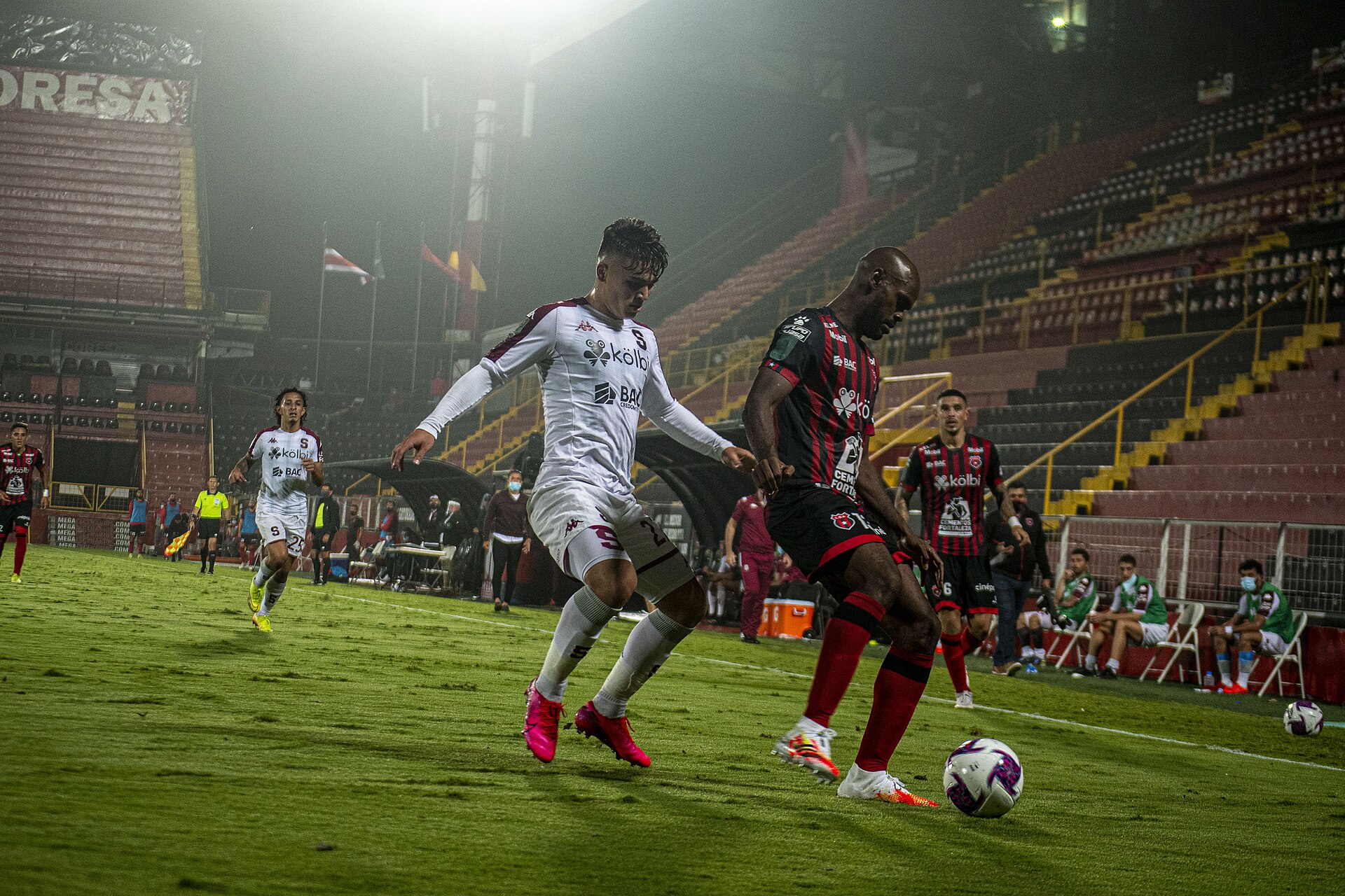 Two players contest the ball on the pitch at the Morera Soto at night, one in a white shirt and one in a red and black Alajuelense shirt