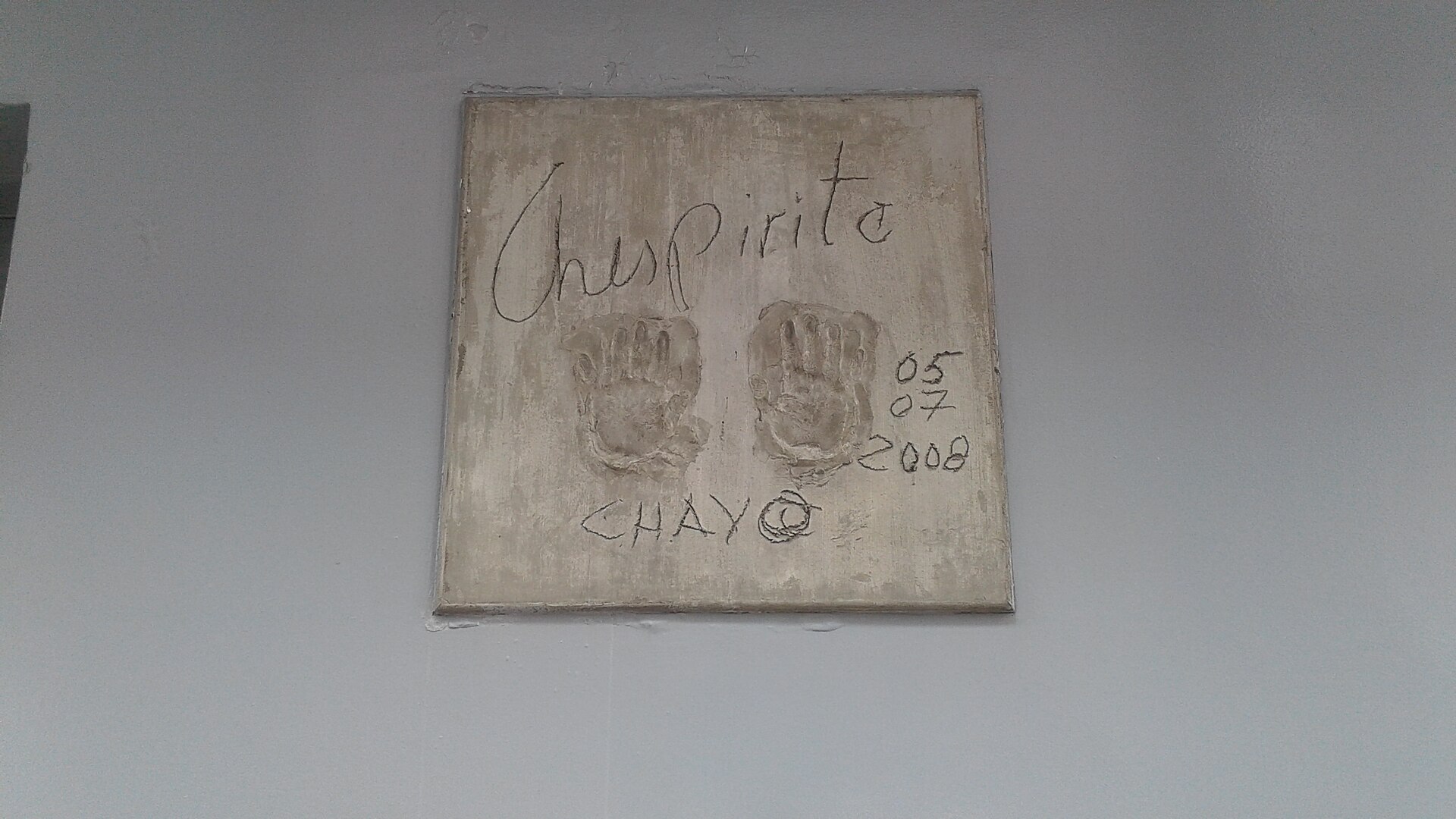 A square cement plaque on a grey wall with two handprints, the handwritten words Chespirito and Chavo, and the date 05 07 2008